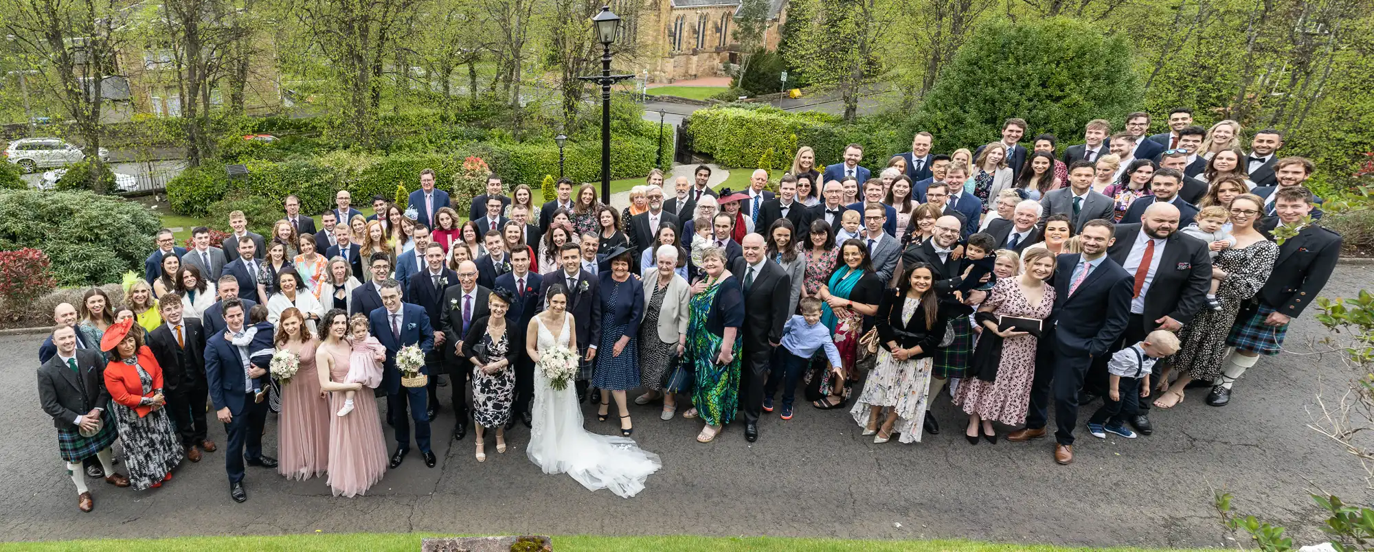Large group of people pose outdoors for a formal event, with trees and a building in the background.