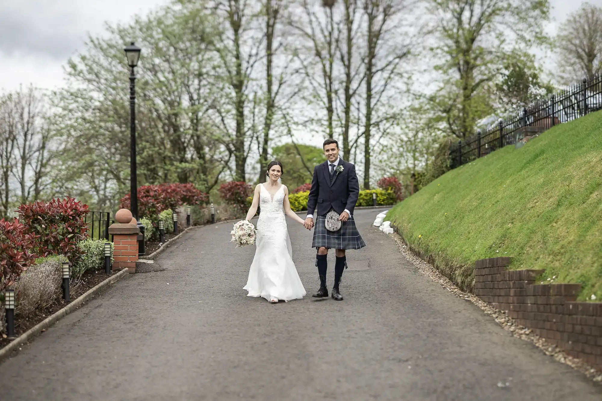 Bride and groom walking hand-in-hand on a paved path, surrounded by greenery and trees, with the groom in a kilt and the bride in a white gown.