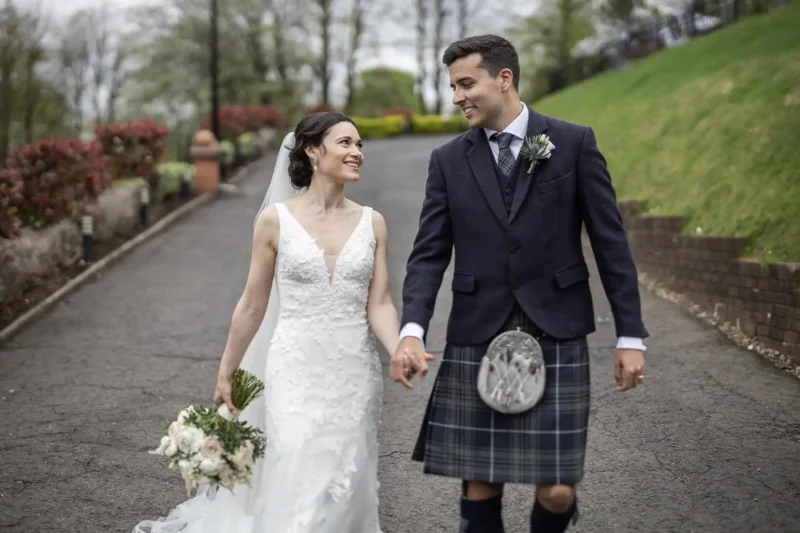 Sherbrooke Castle Hotel wedding photos: Bride in white dress and groom in a kilt walking on a tree-lined path, holding hands and smiling at each other.