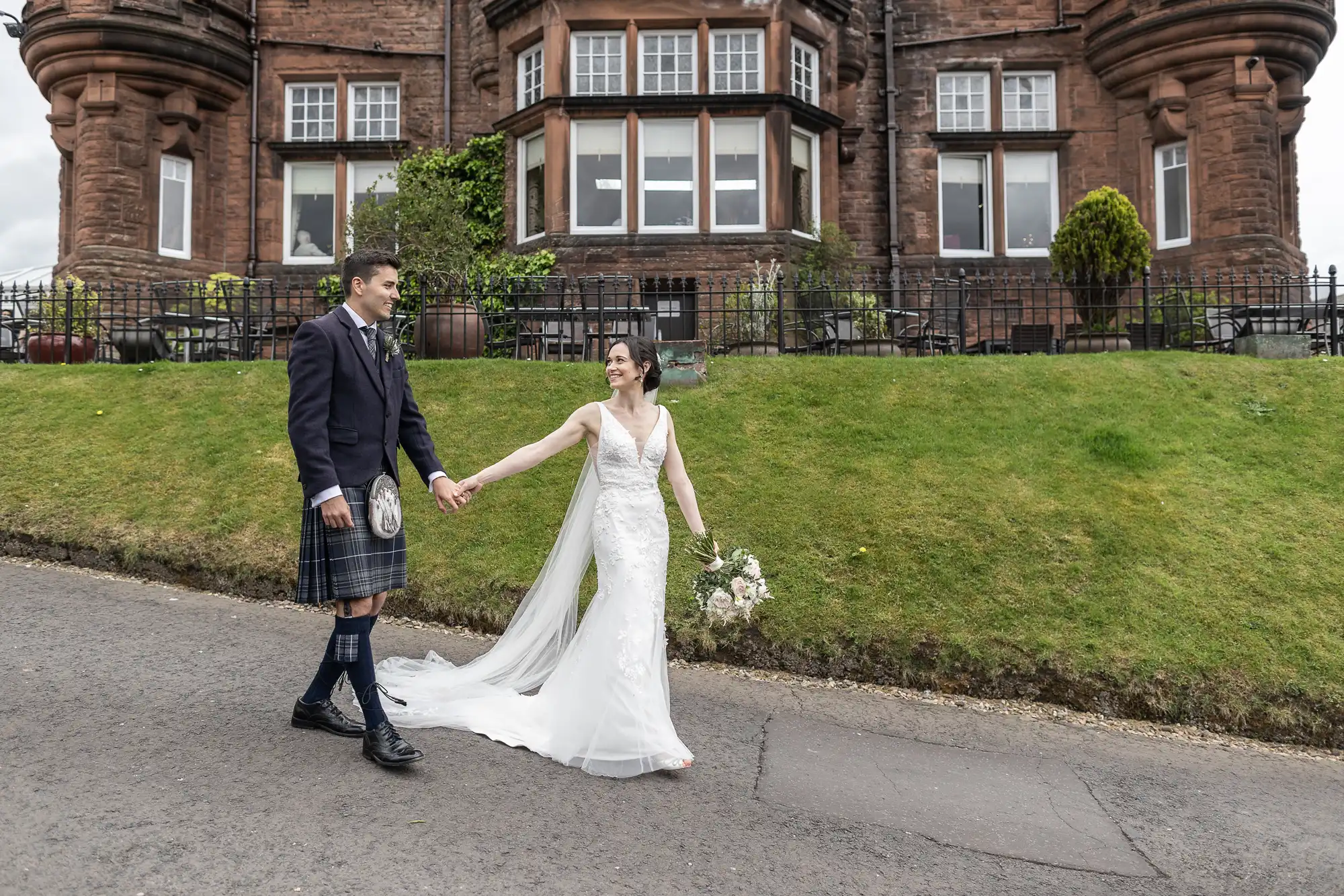 Bride and groom holding hands, walking in front of a historic stone building with grass and plants. The groom wears a kilt and the bride, a white gown.