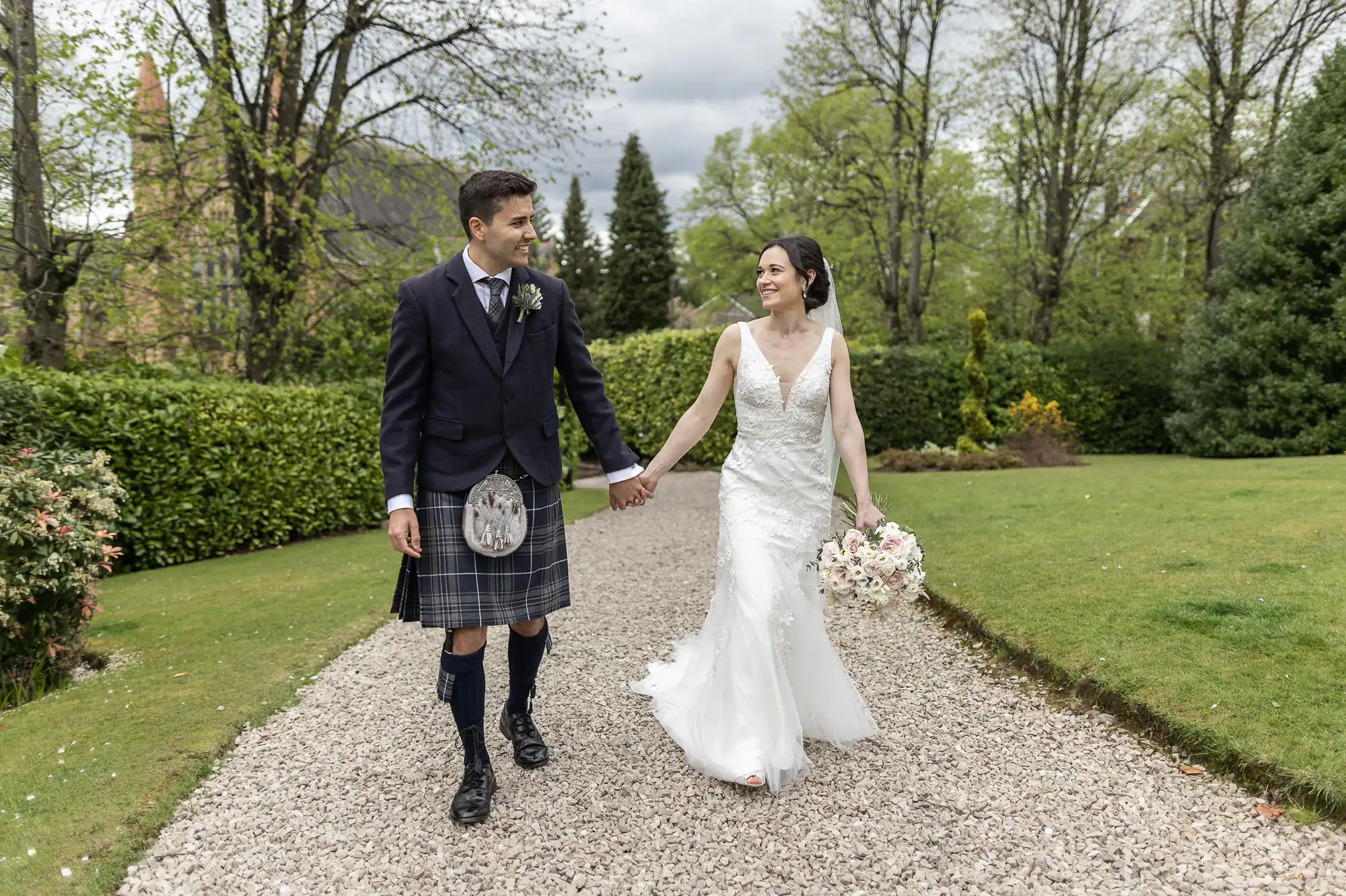 A couple walks hand in hand on a gravel path surrounded by greenery; the man wears a kilt, and the woman is in a white dress holding a bouquet.