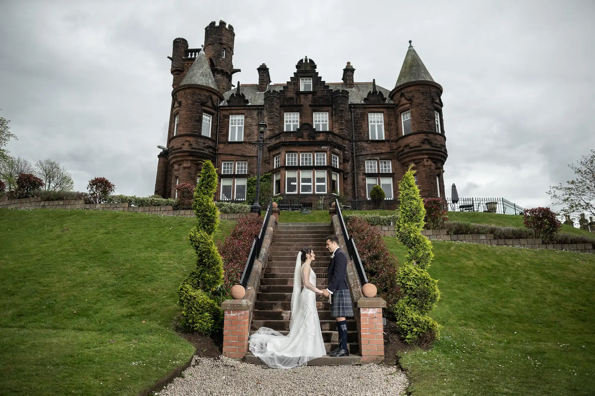 A bride and groom stand on steps in front of a large historic stone building with cones and turrets, surrounded by greenery and a cloudy sky.
