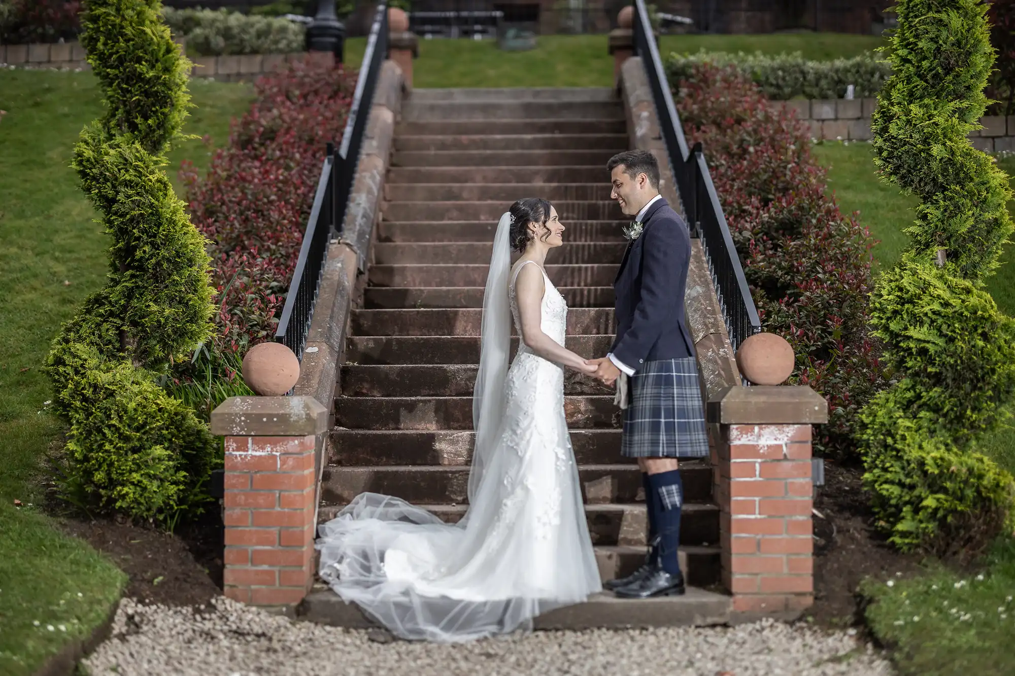 A bride and groom stand holding hands in front of brick steps, surrounded by greenery. The groom wears a kilt, and the bride is in a white dress with a veil.