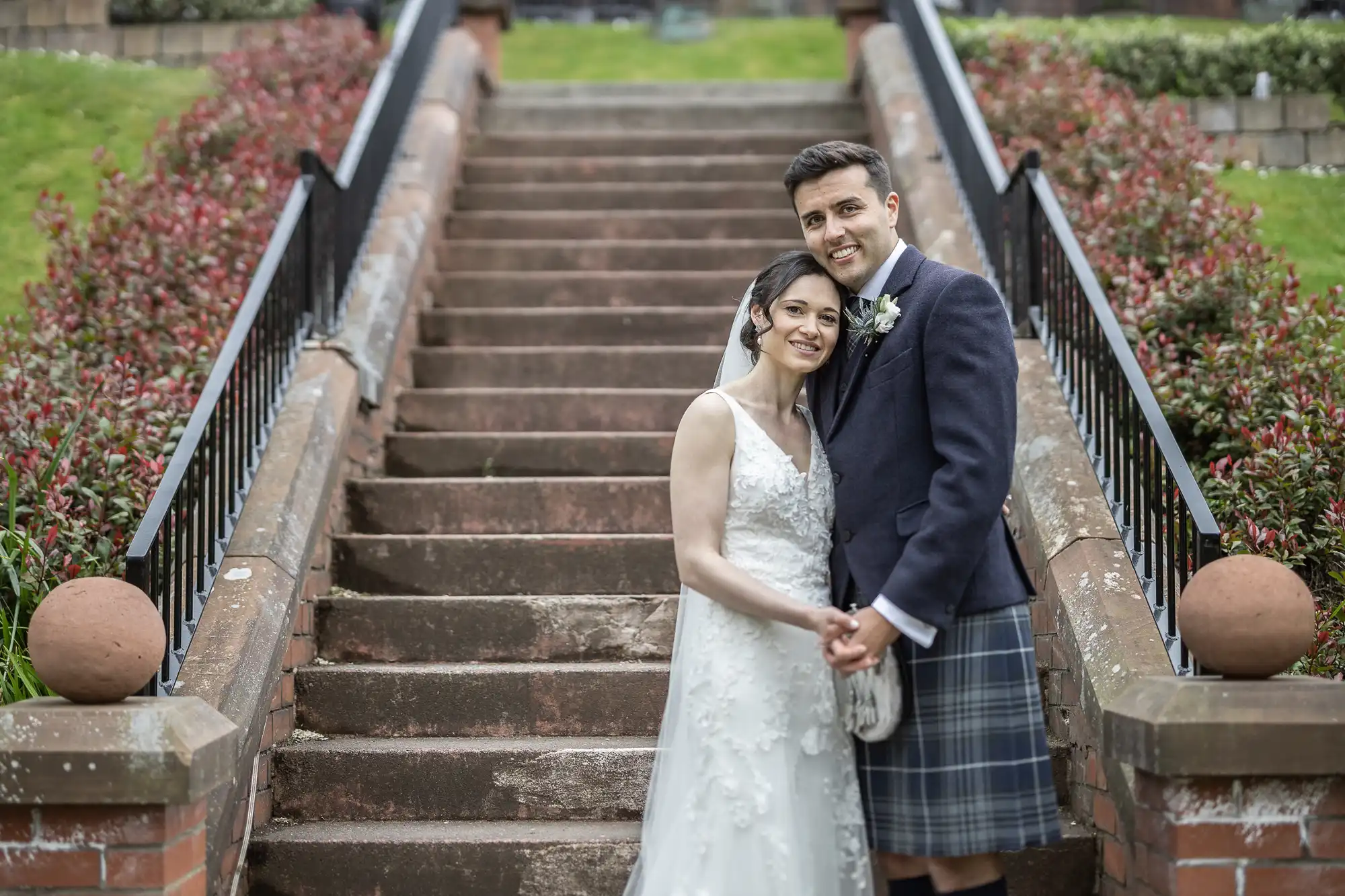 A couple in wedding attire poses on outdoor stone steps. The woman wears a white dress and veil, and the man wears a dark jacket and kilt.