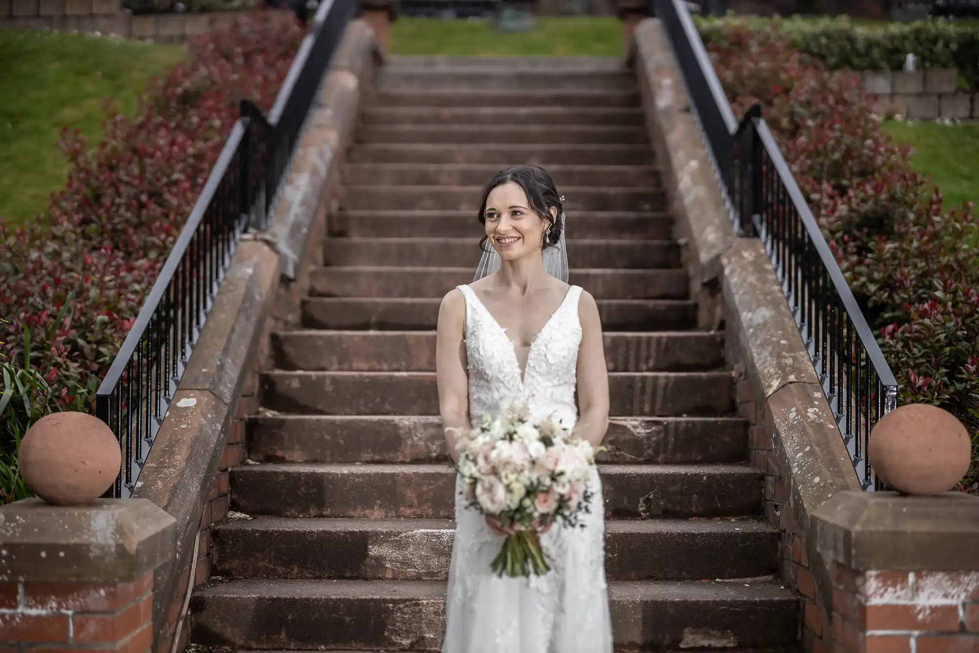 Bride in a white gown holding a bouquet stands on outdoor steps, smiling.