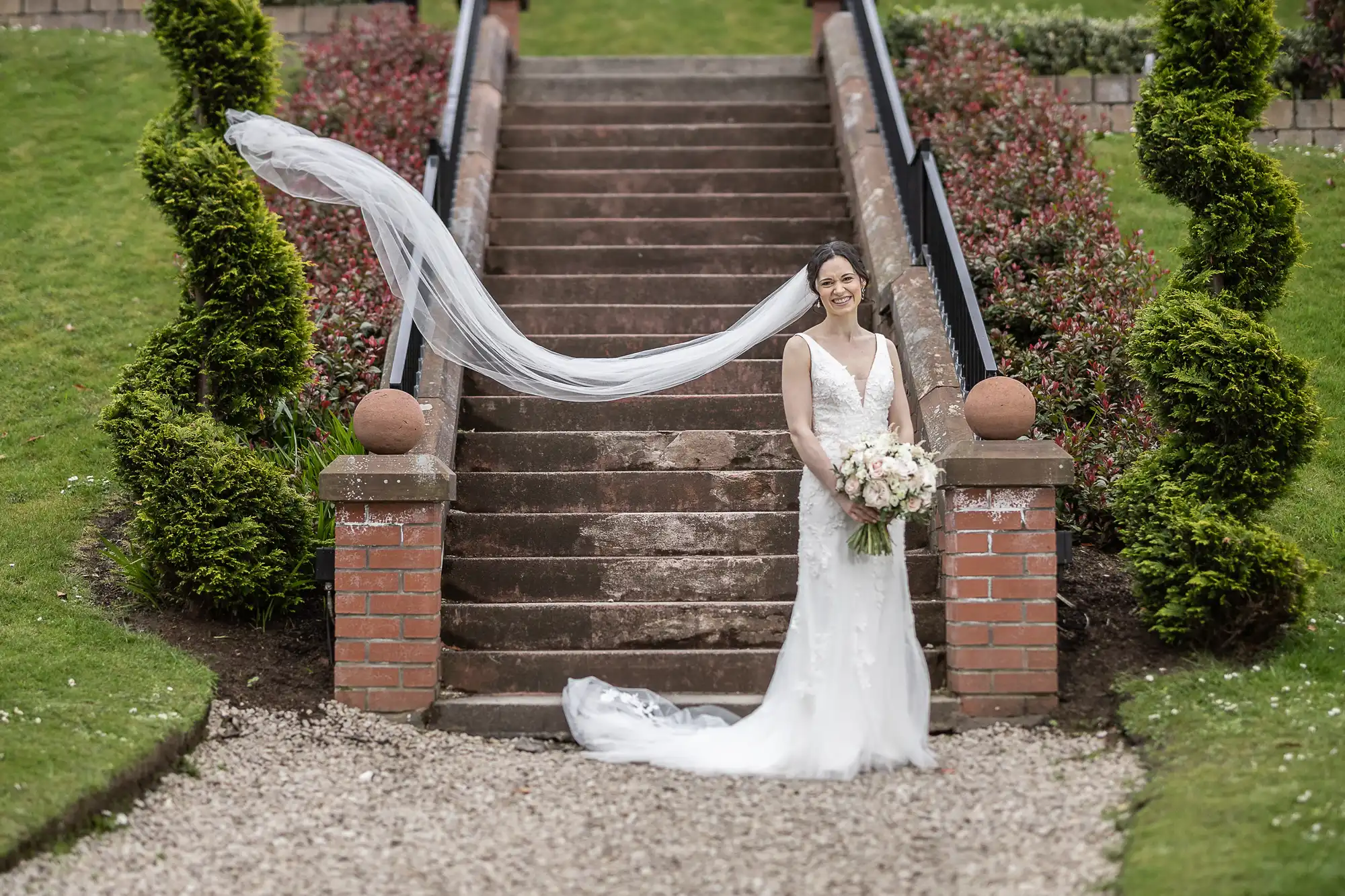 A bride in a white dress stands at the bottom of stone steps in a garden, holding a bouquet, with her veil blowing behind her.