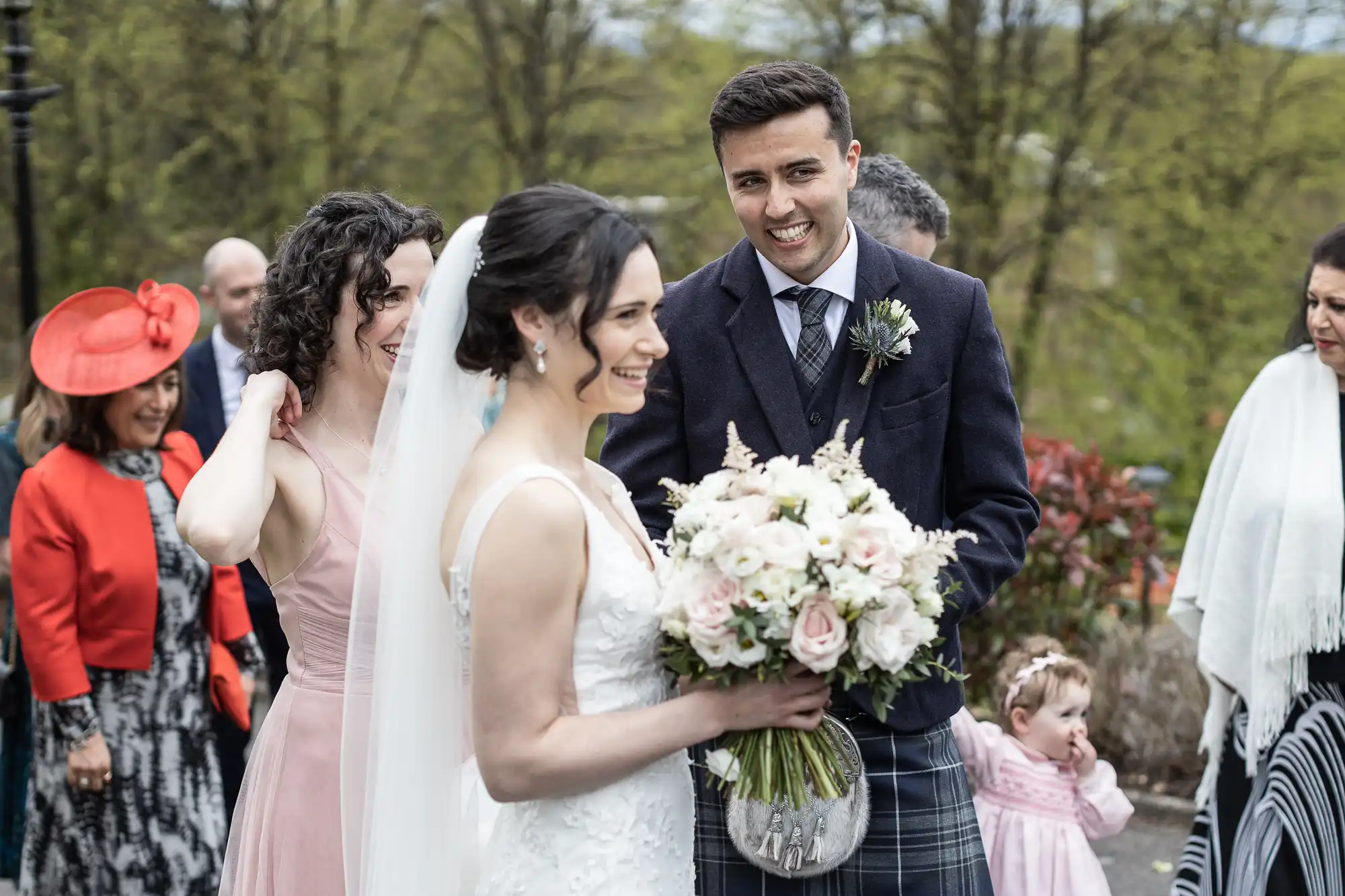 A bride and groom smile while holding a bouquet. They are surrounded by guests outdoors. A woman in red and a child in pink are visible in the background.