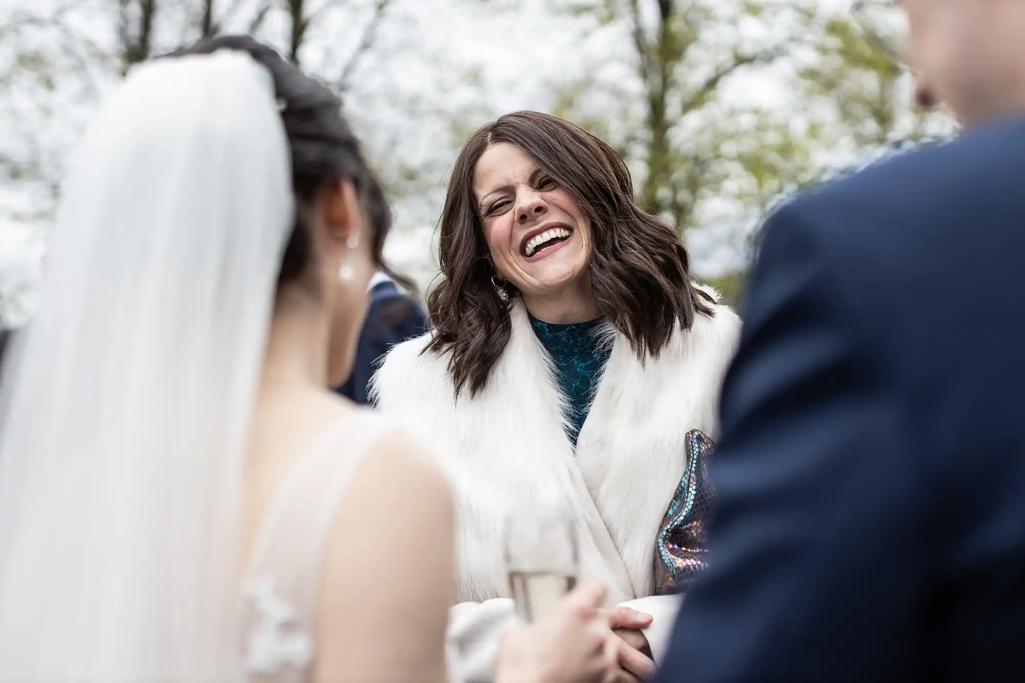 Woman in a white fur coat laughs joyfully while standing with a bride in a white dress and veil in an outdoor setting.