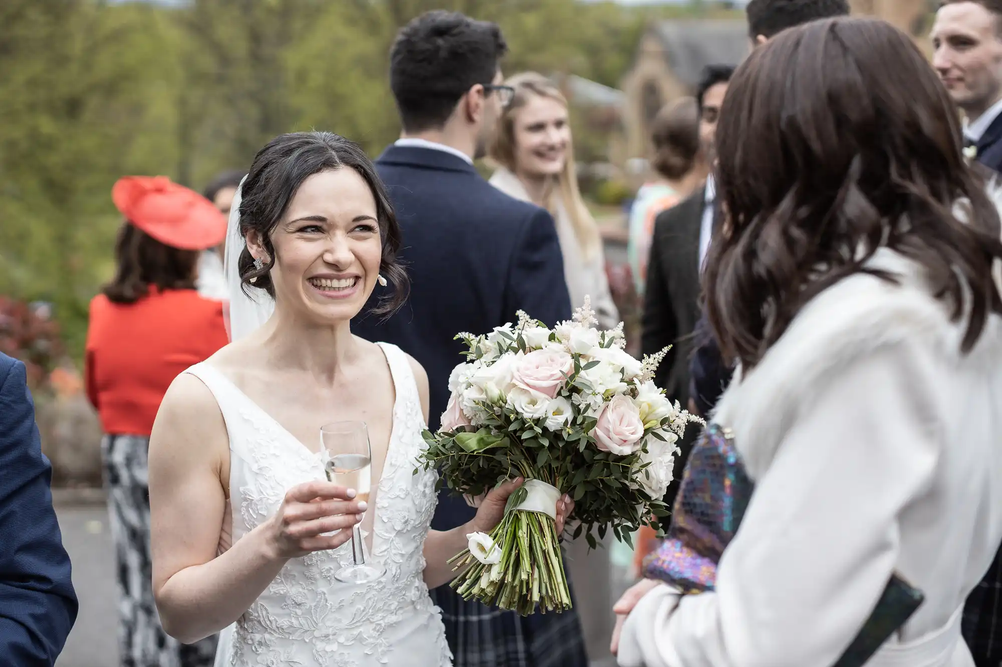 A bride in a white dress smiles while holding a bouquet and a glass, talking to a guest at an outdoor gathering. Other people in formal attire are in the background.