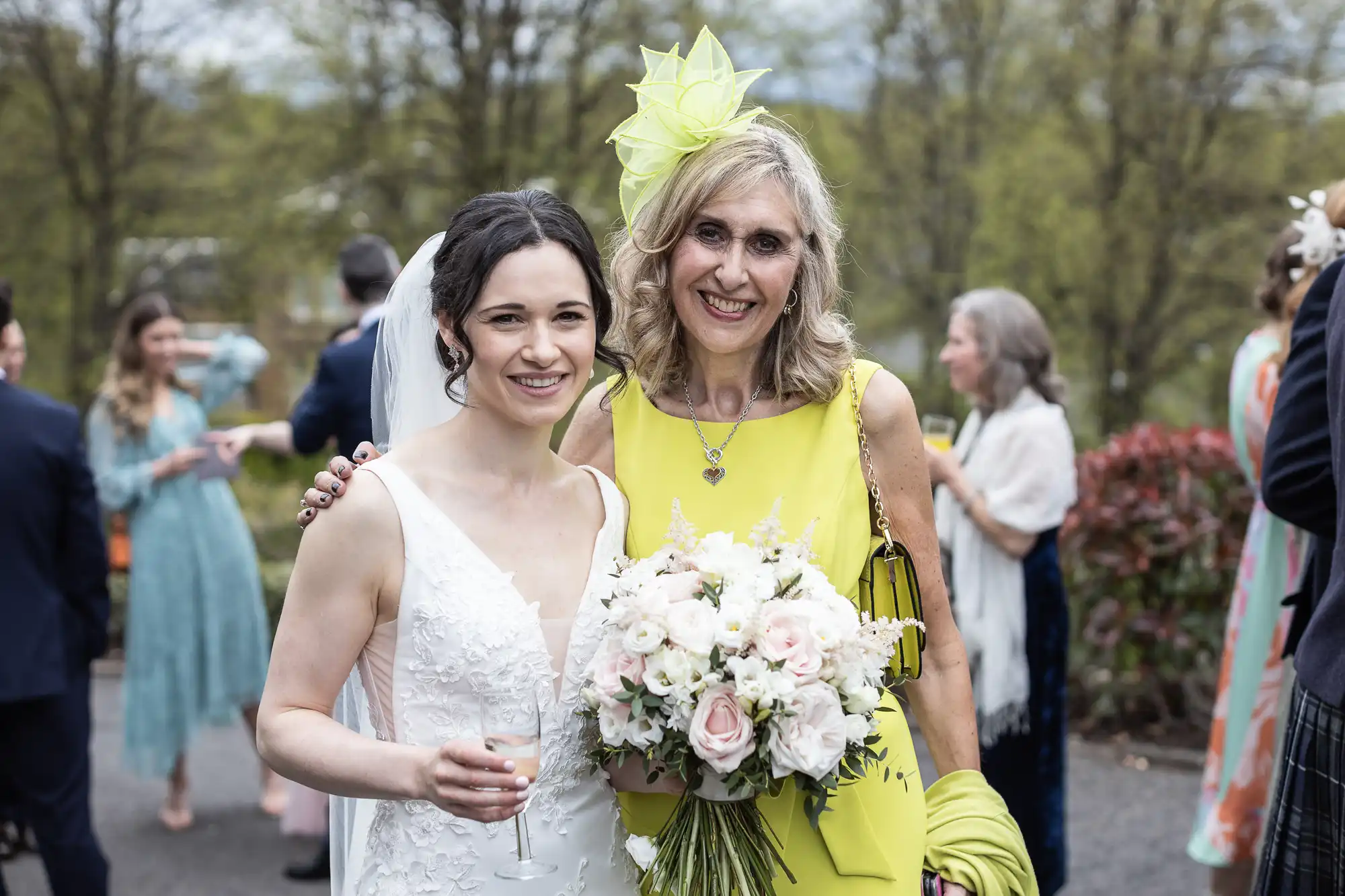 Two women smiling, dressed in wedding attire. One is in a white gown holding a bouquet, the other in a yellow outfit with a matching fascinator. People in background with trees visible.