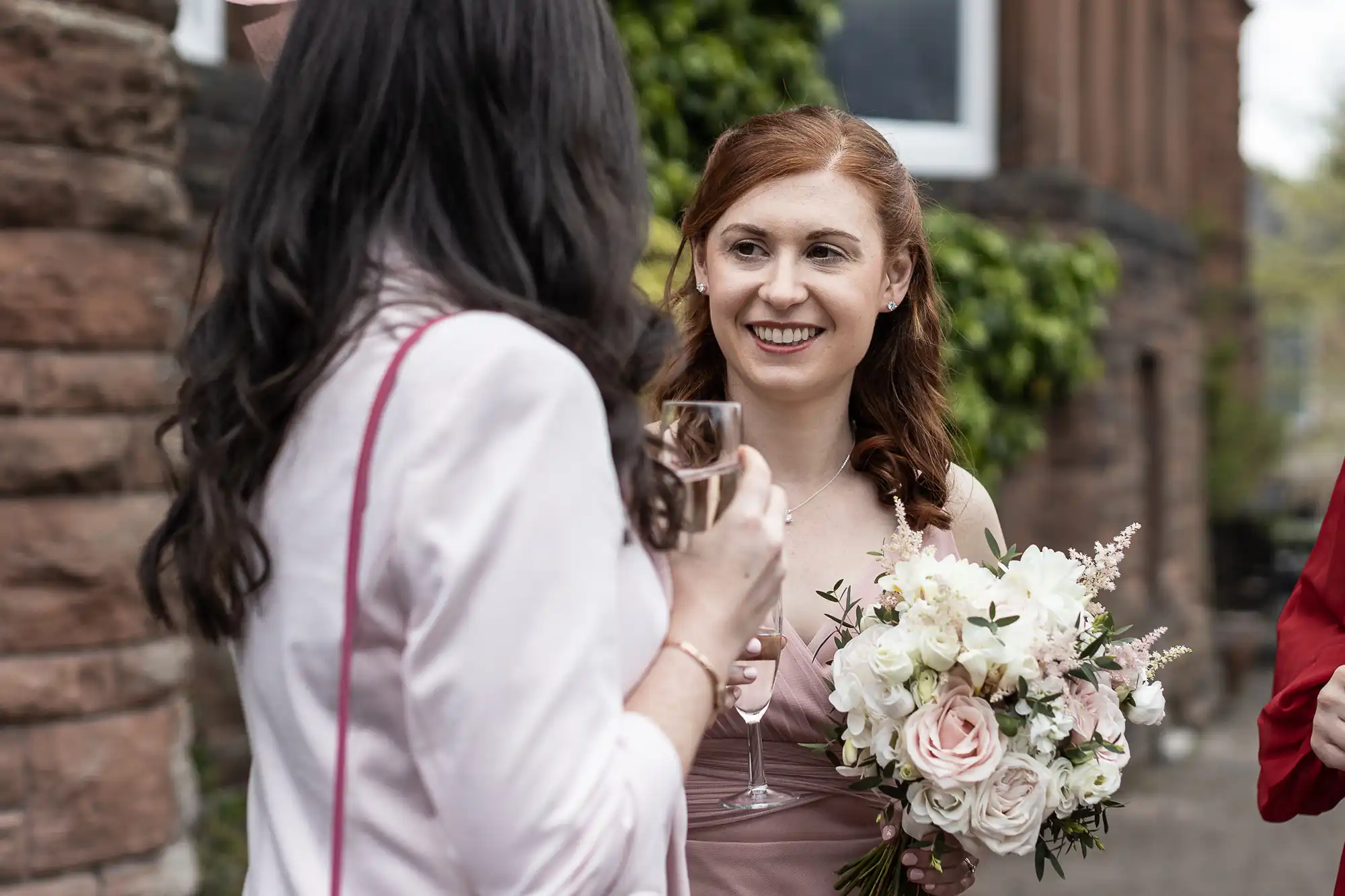 Two women at an outdoor event. One holds a wedding bouquet and smiles at the other, who is holding a drink. Both wear pink attire, and there are green plants and a brick wall in the background.