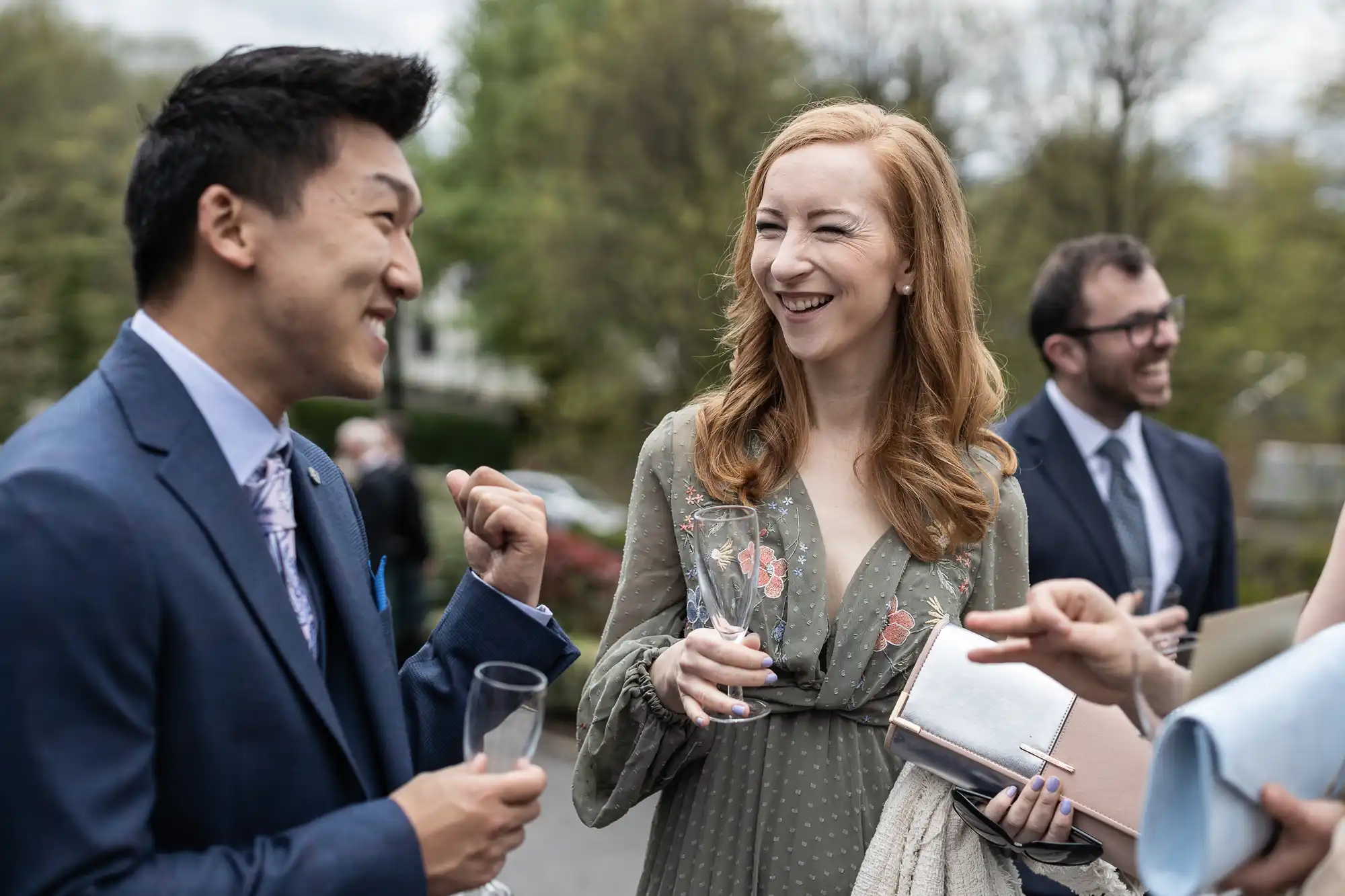 People in formal attire are smiling and talking outdoors, holding glasses. Trees are visible in the background.