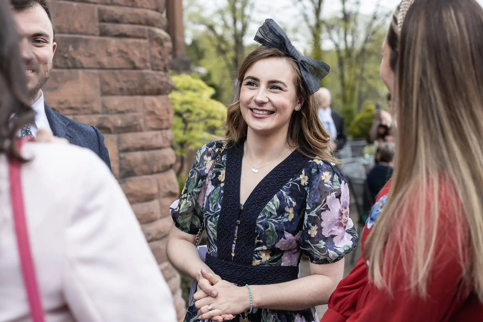 A woman in a floral dress and headband smiles while standing outside, engaging in conversation with three people.