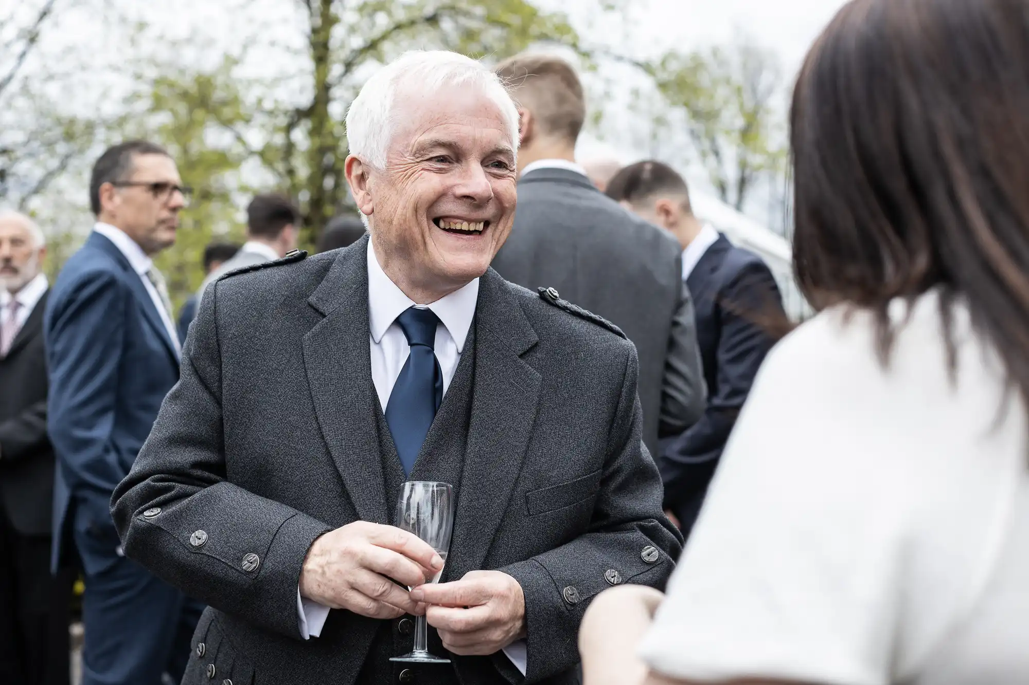 An elderly man in a suit smiles and holds a champagne glass while talking to a woman at an outdoor event with other people in the background.