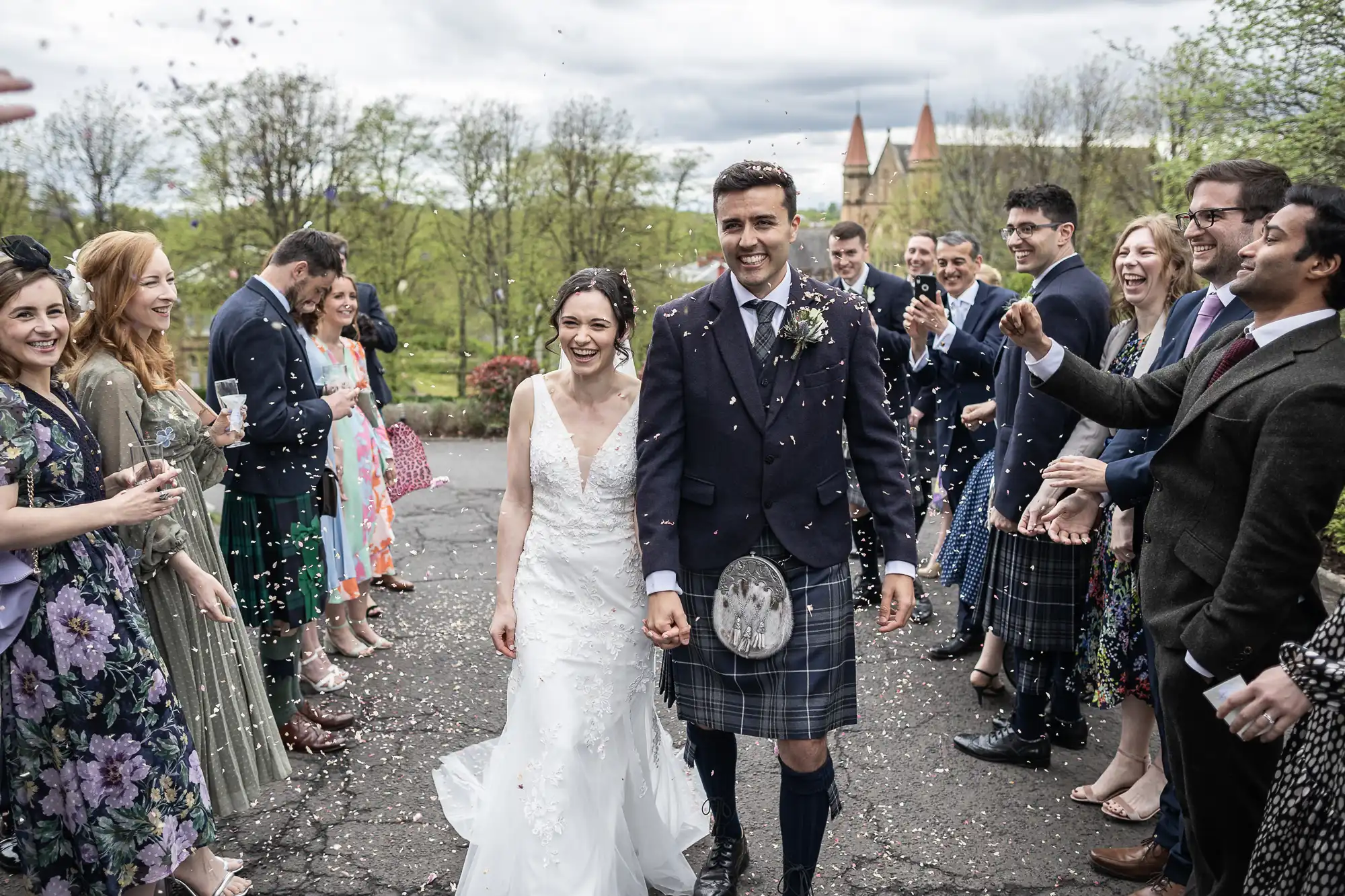 A bride and groom walk through a crowd of people throwing confetti. The groom wears a kilt, and the bride is in a lace wedding dress. Trees and a building are visible in the background.