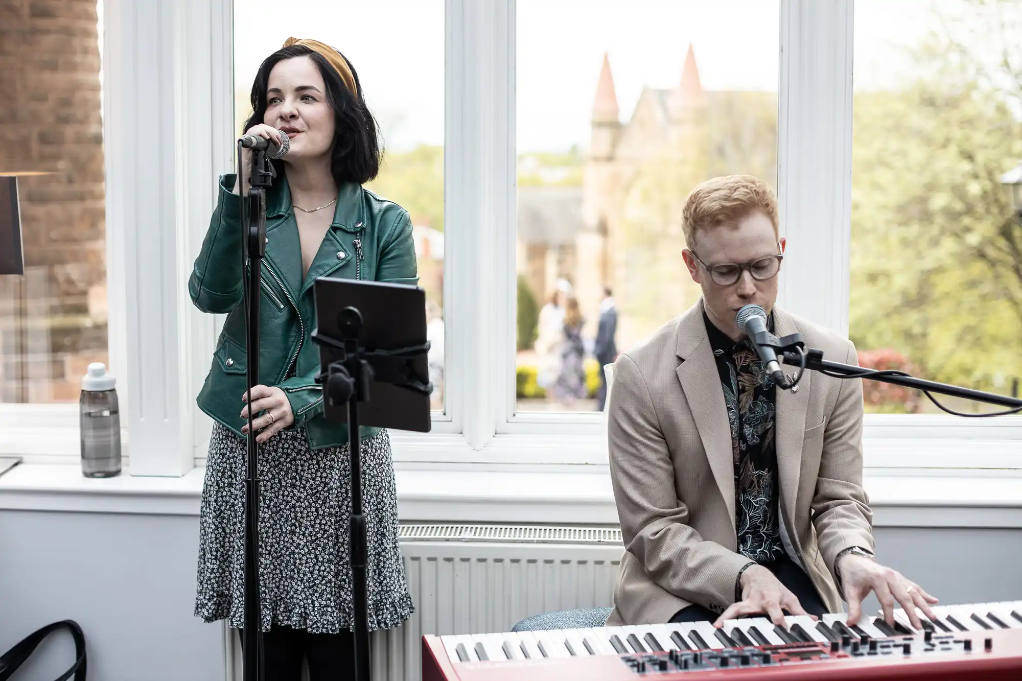 A woman sings into a microphone while a man plays a keyboard. They perform in a room with large windows showing a view of trees and a brick building outside.