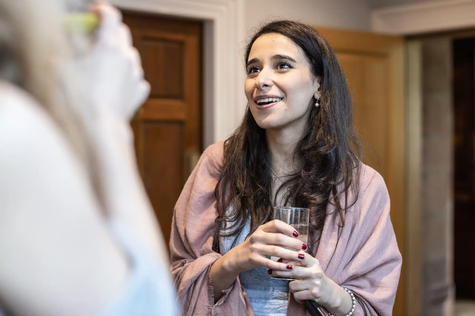 Woman with long hair holds a glass and listens attentively indoors, wearing a pink shawl and smiling slightly.