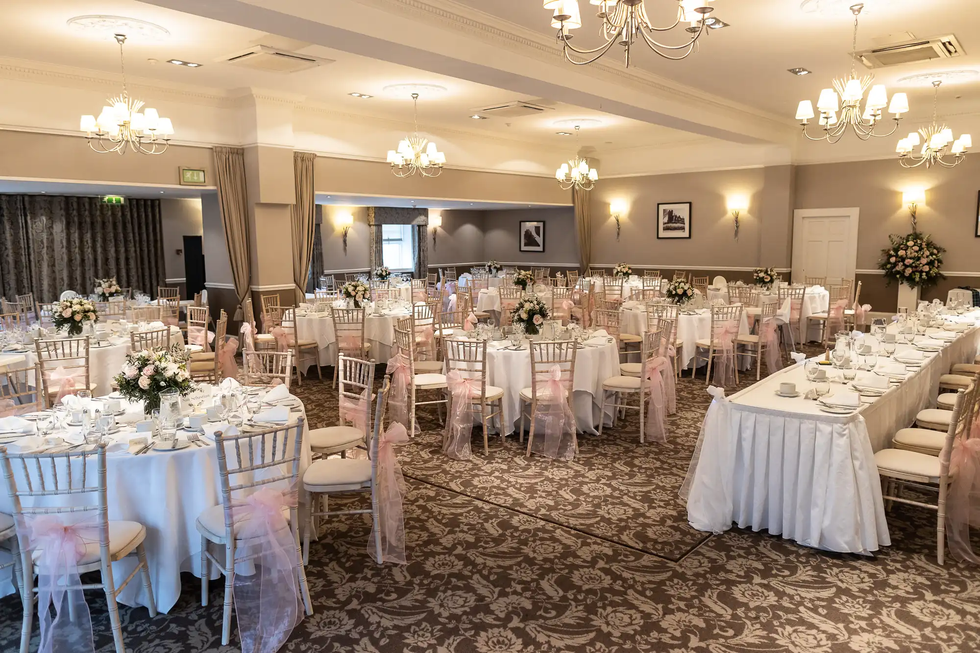 Elegant banquet hall set for an event with round tables, white tablecloths, floral centerpieces, and decorative chair covers. Chandeliers hang from the ceiling.
