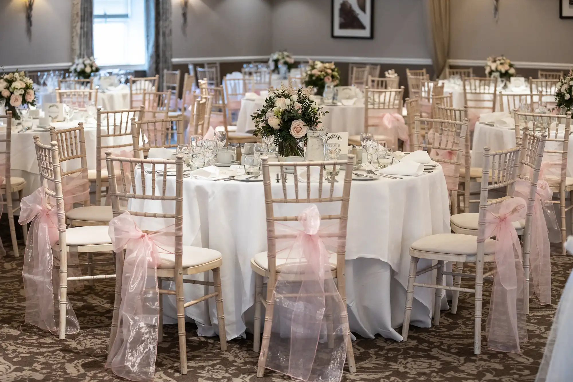 Elegant wedding reception setup with round tables, white tablecloths, and floral centerpieces. Chairs have pink ribbon accents.