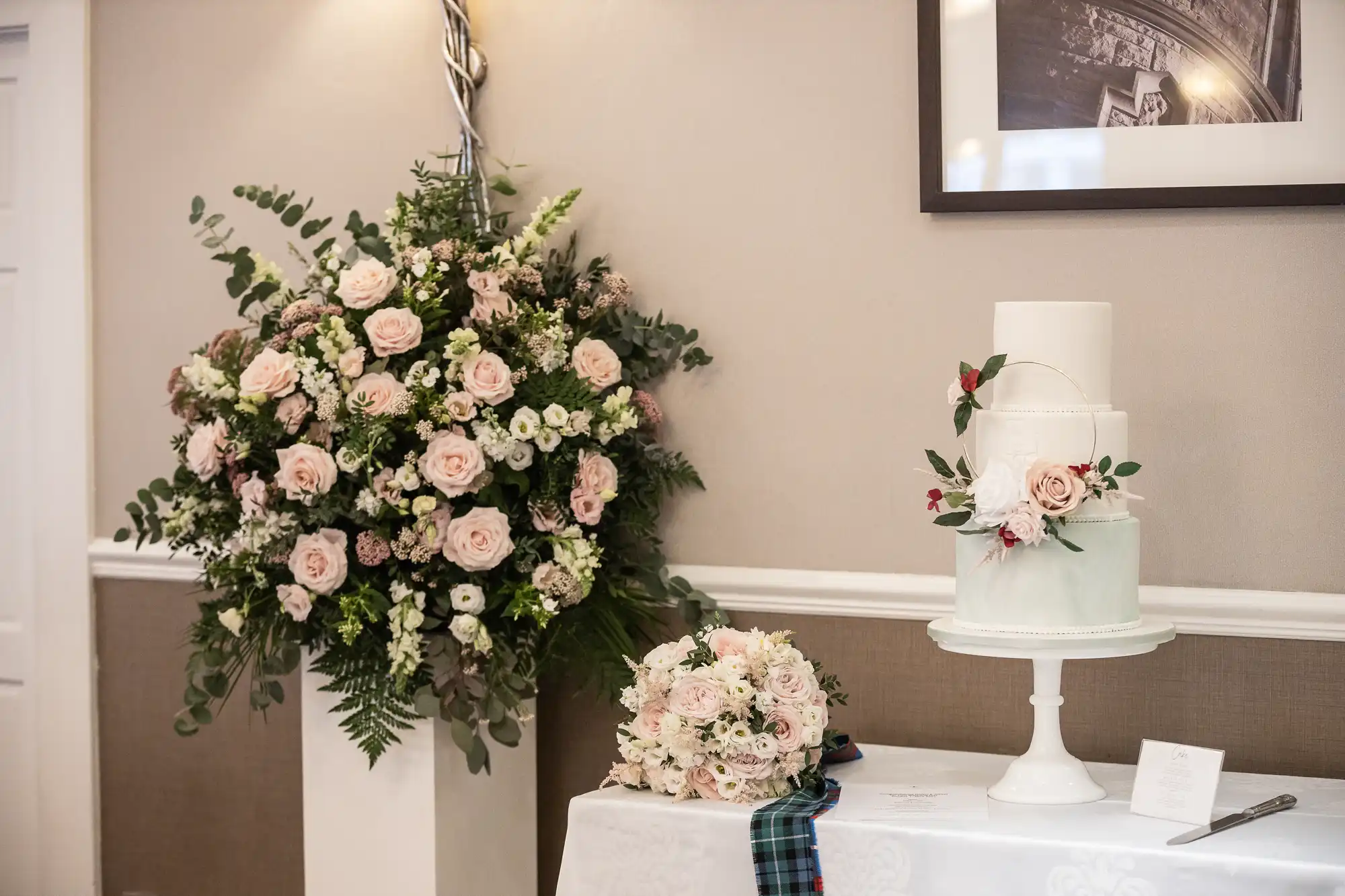 A floral arrangement and a three-tiered wedding cake with floral decorations are on a table. There's a bouquet and a tartan ribbon nearby. A framed picture hangs on the wall.