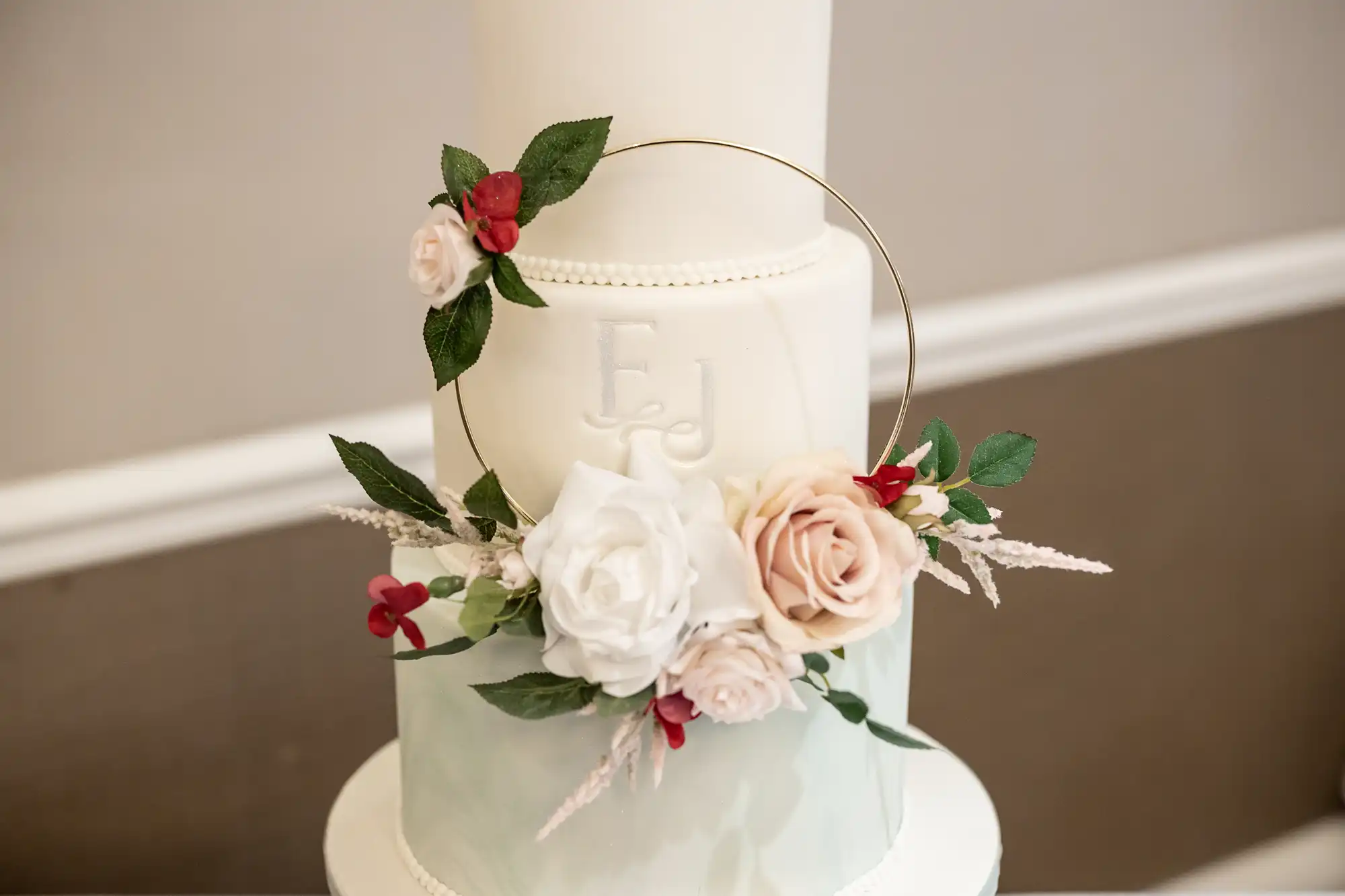 Two-tiered white cake with initials "EJ" features pink and white roses with greenery and a gold ring decoration.