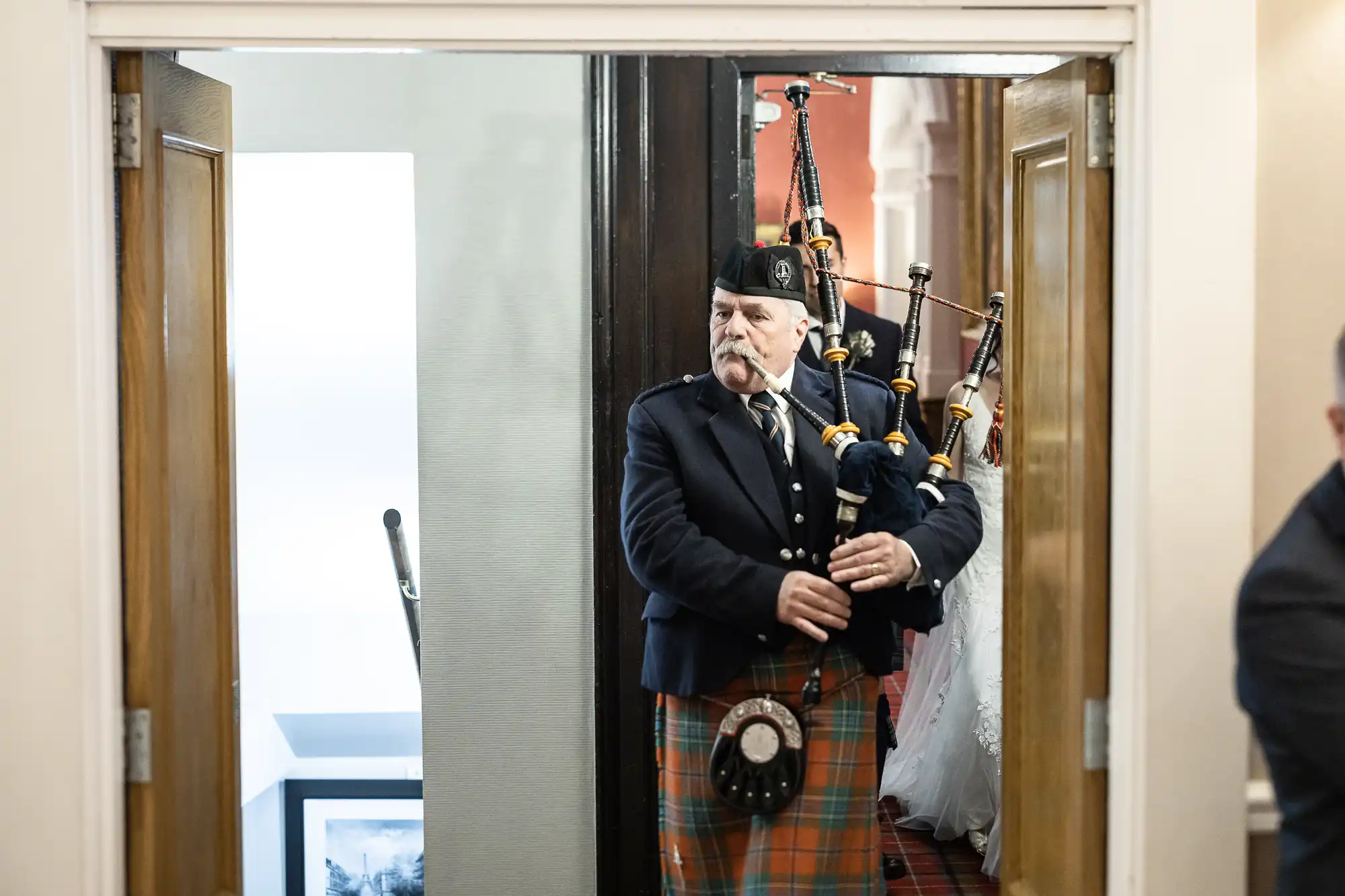 A man in traditional Scottish attire plays bagpipes while walking through a doorway.