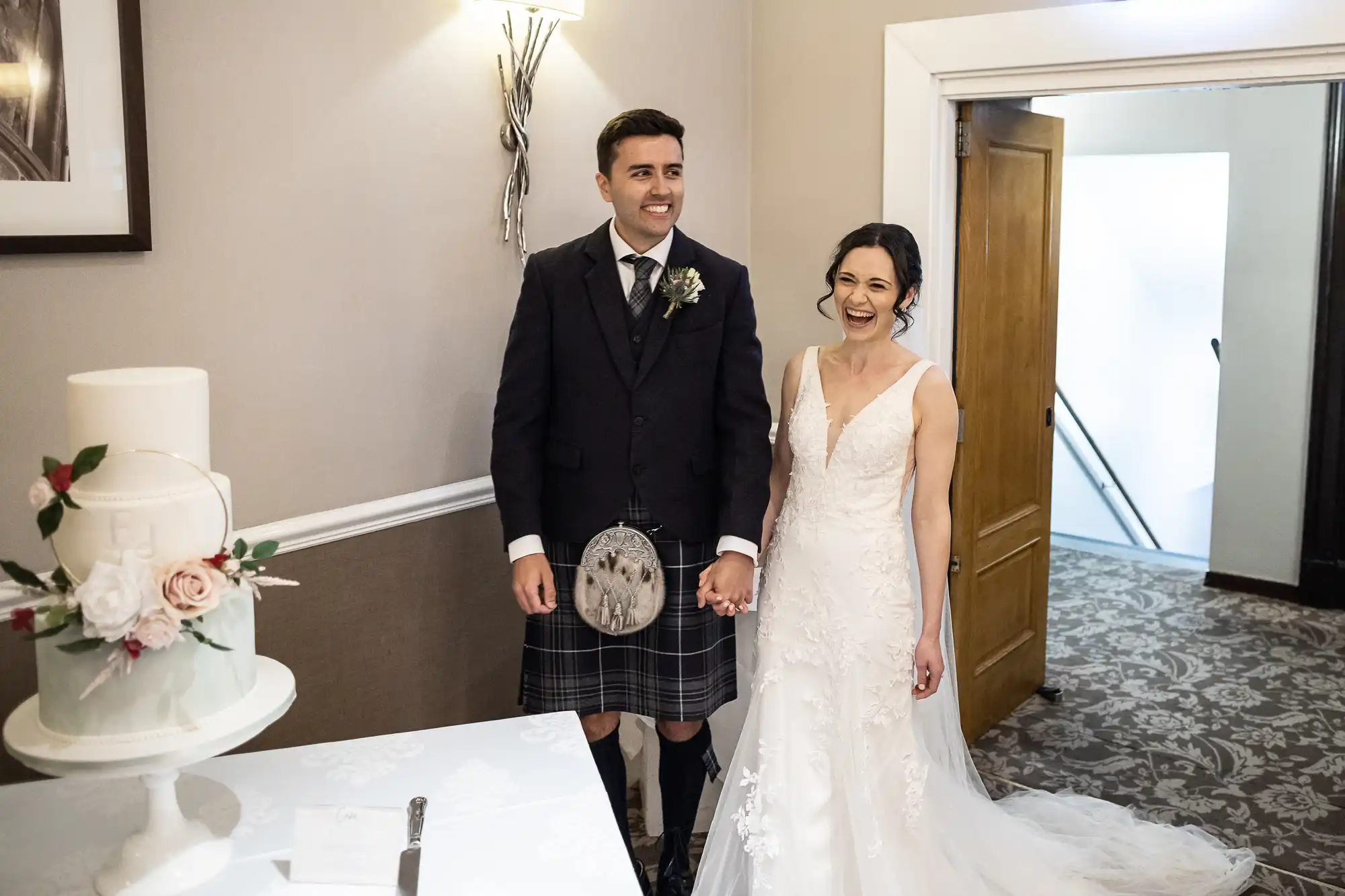A couple in wedding attire stands smiling in a room. The man wears a kilt, and the woman is in a white gown. A white cake with flowers is on a table beside them.