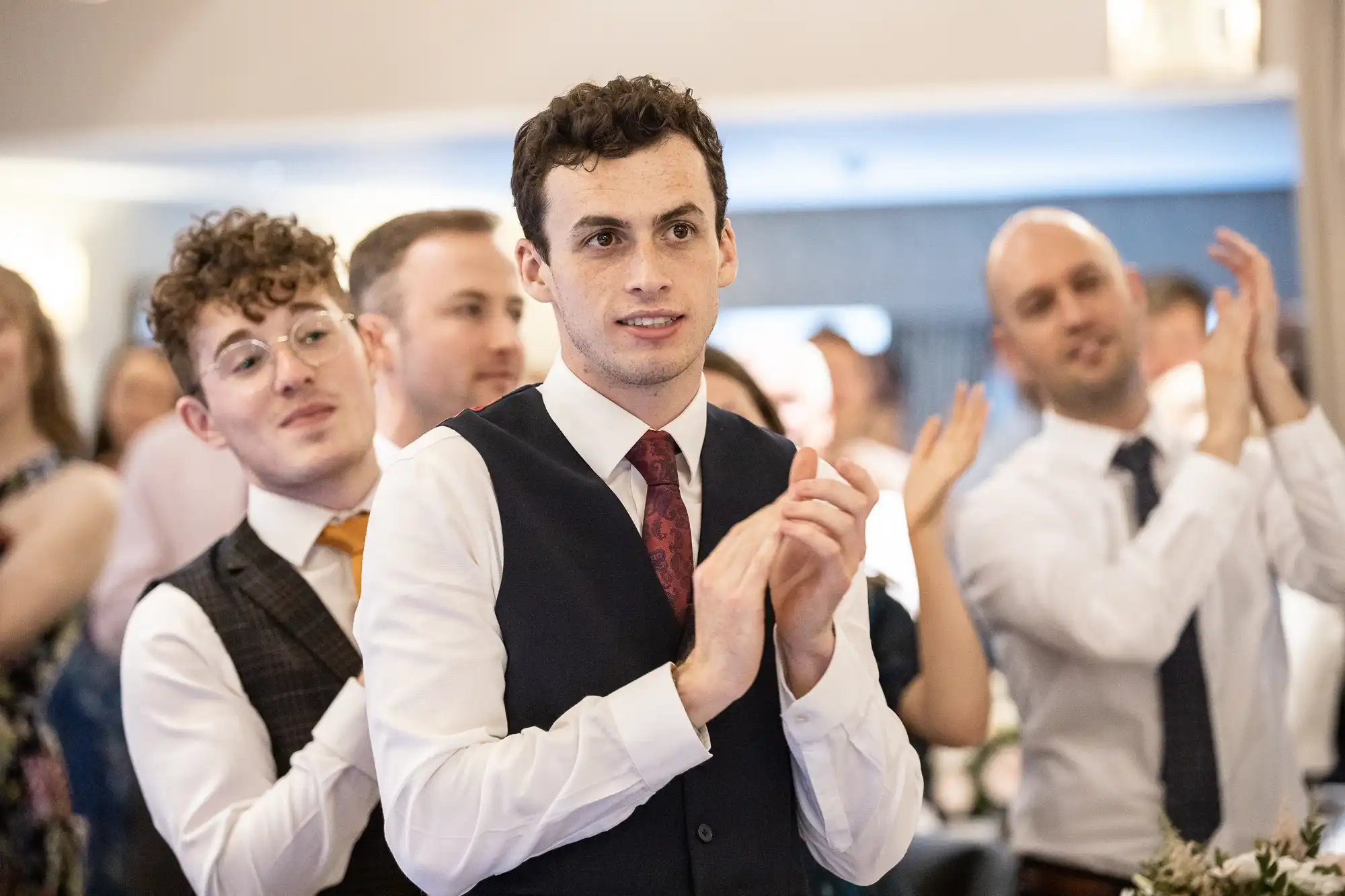A group of people in formal attire clap at an indoor event, with a focus on a man in a vest and tie.