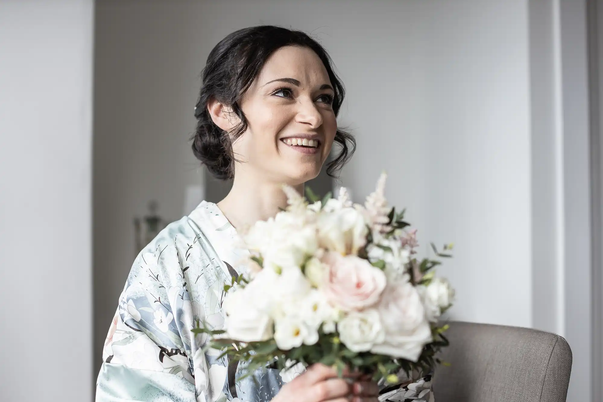 Woman smiling, wearing a floral robe, holding a bouquet of white and pink flowers indoors.