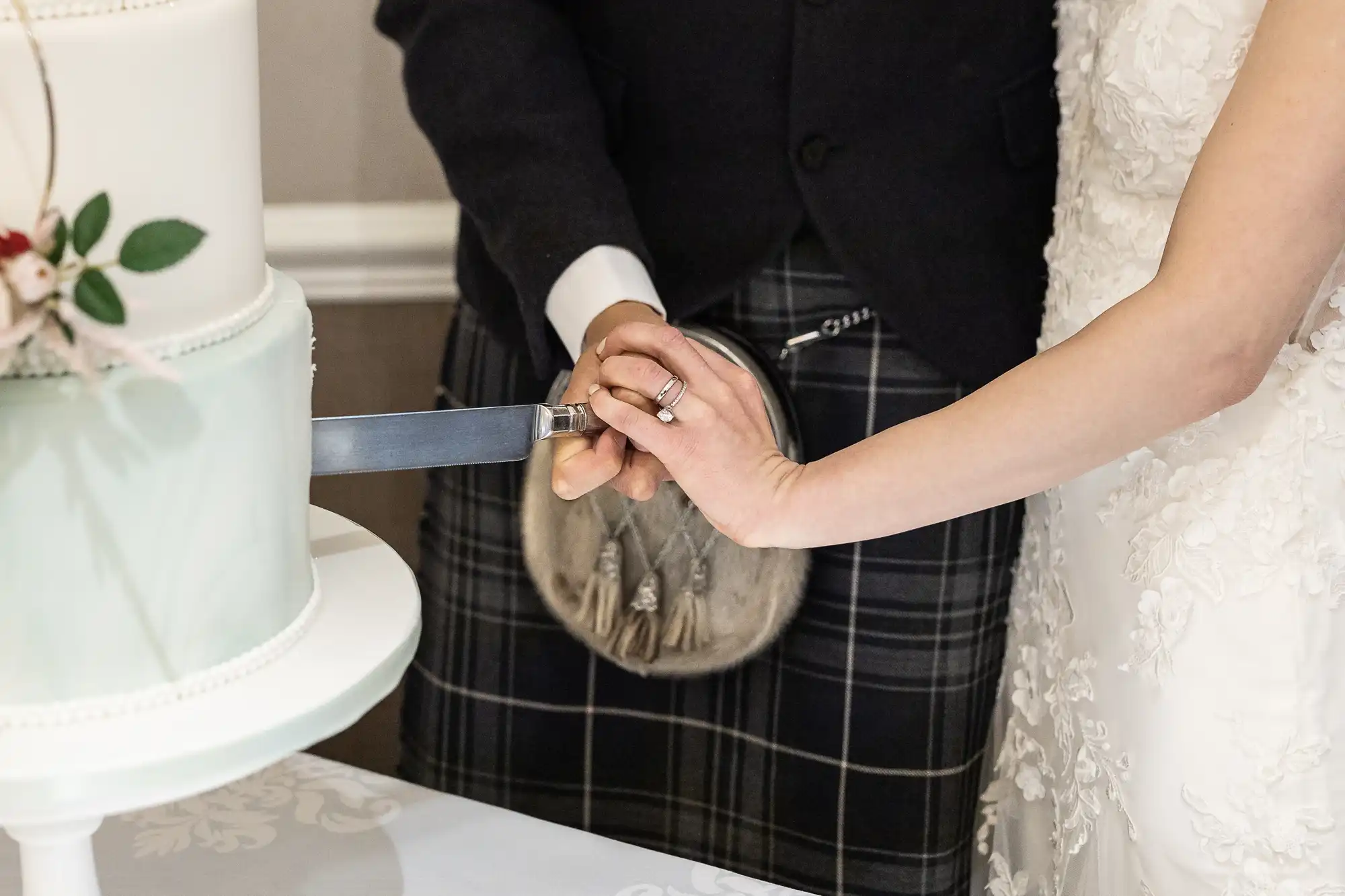 A couple cuts a wedding cake together. One wears a suit with a kilt and sporran, the other wears a white wedding dress.