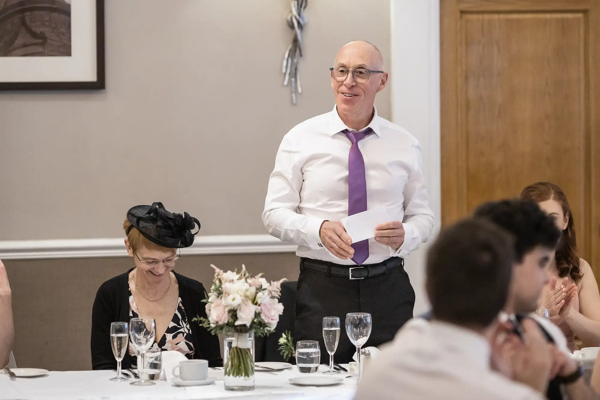 An older man in a white shirt and purple tie stands holding a note, speaking at a table during a formal event. A woman sits nearby, and flowers and tableware are visible.