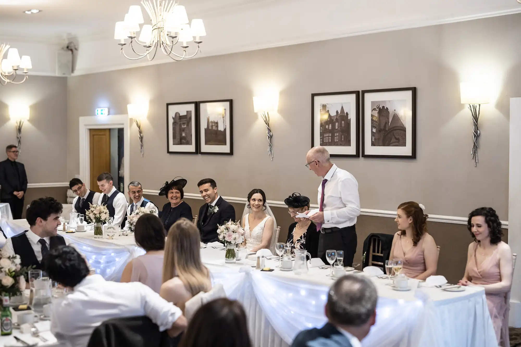 A man stands and speaks at a long table during a formal event. Seated guests, some in formal attire, listen and smile. The room features framed artwork and decorative lights.