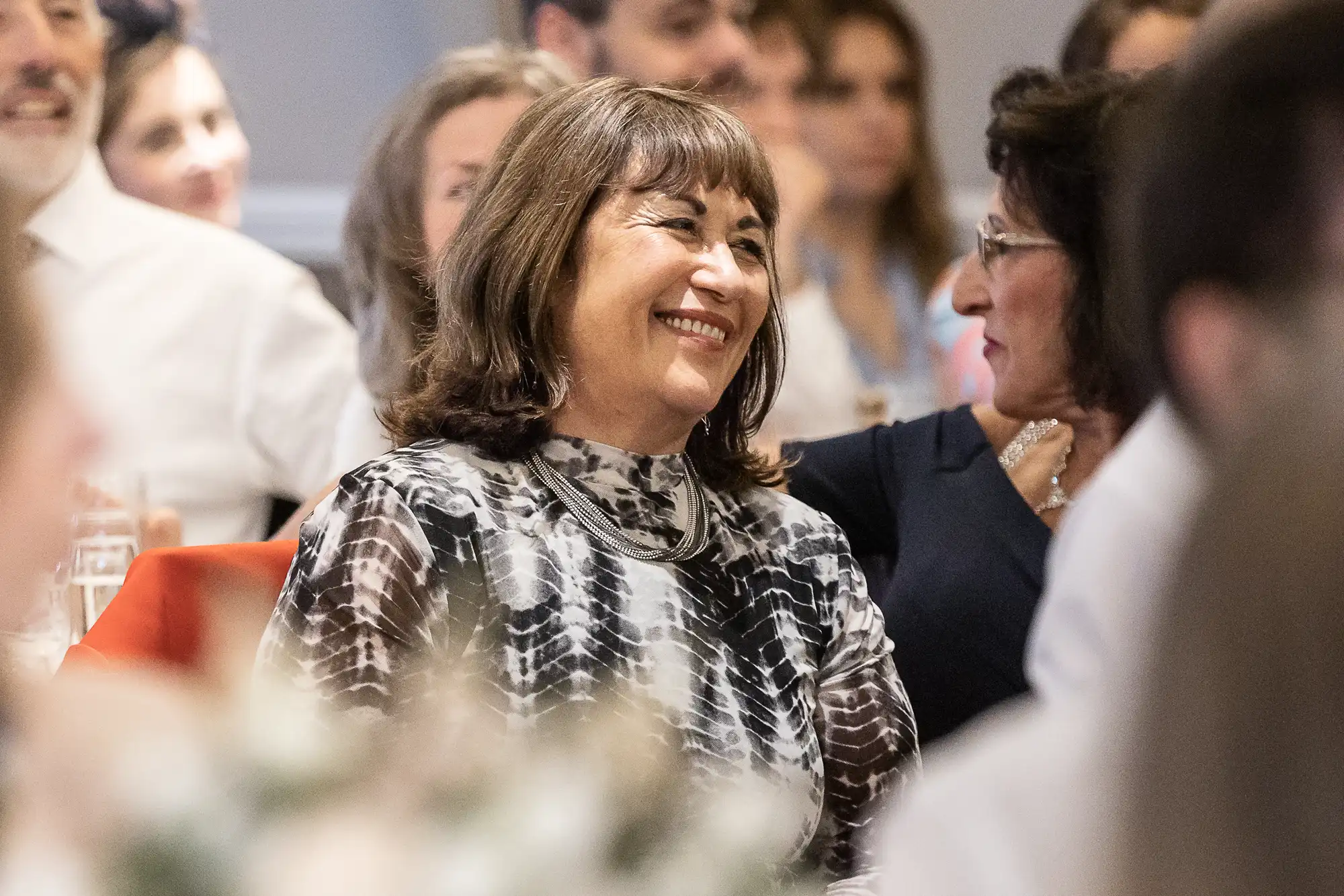 A woman with shoulder-length hair and a patterned top smiles while sitting in a crowd of people.