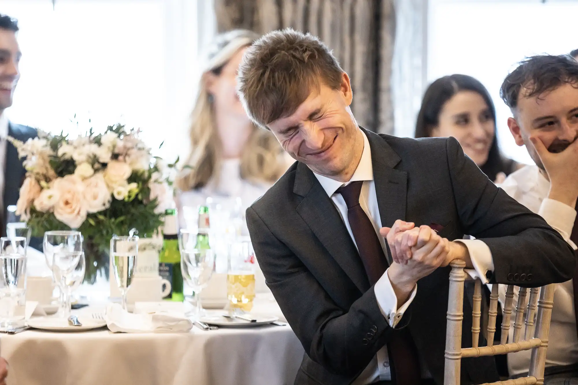 A man in a suit smiling with eyes closed, seated at a table with flowers and drinks, surrounded by other people.