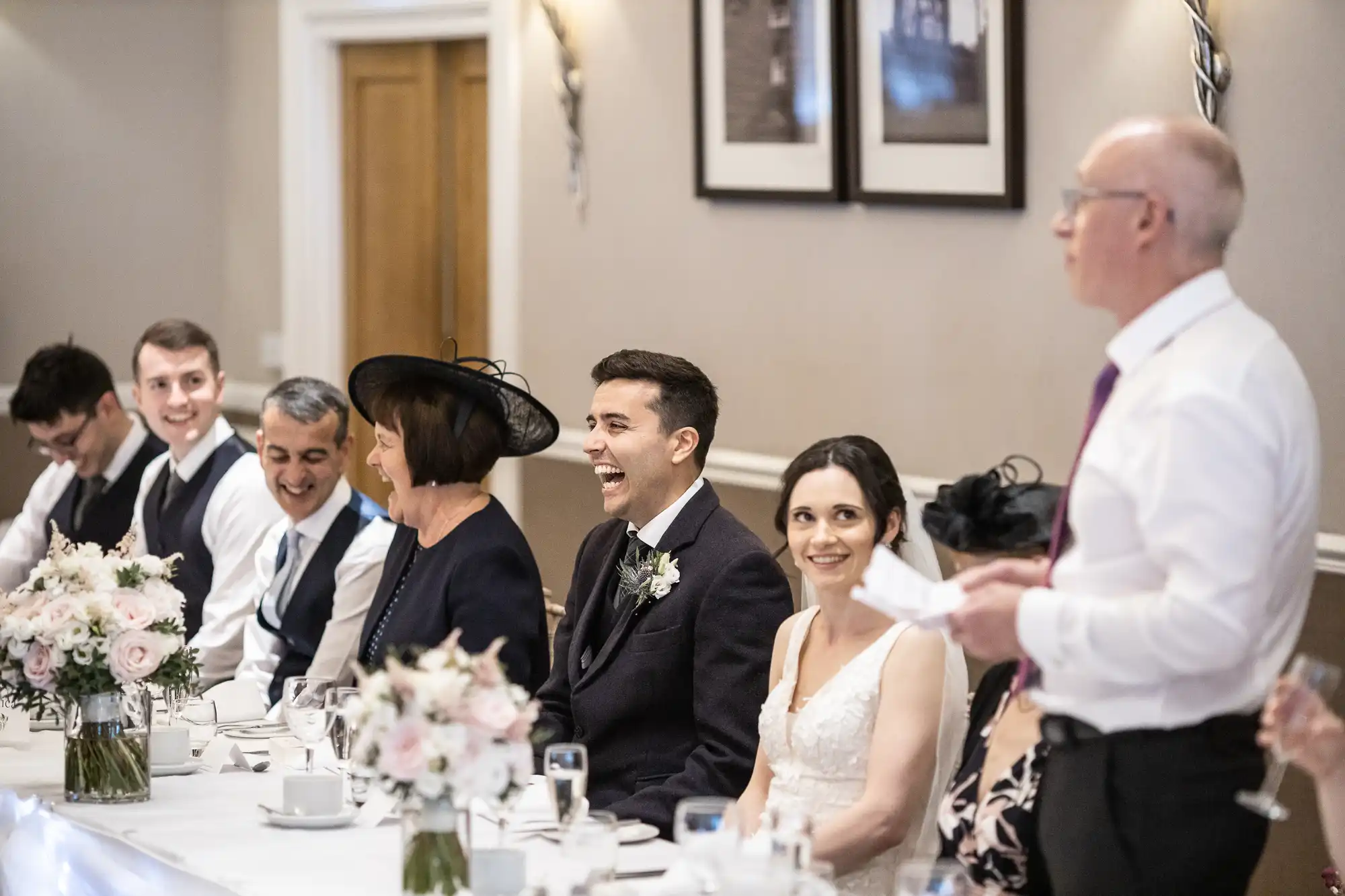 People sitting at a long table, dressed in formal attire. A man in a white shirt stands, speaking, while the others listen. There are floral decorations on the table.