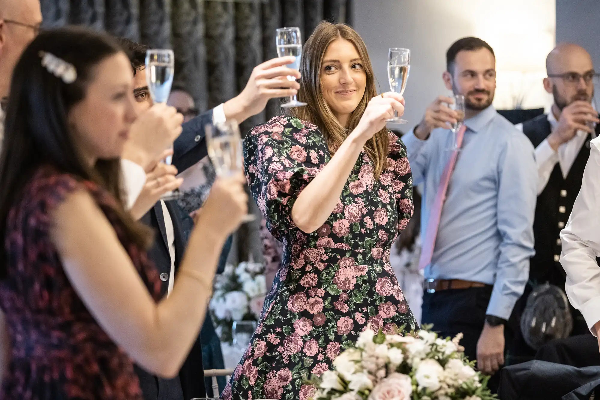 People in formal attire raising glasses in a toast at an event, with floral arrangements on the table.