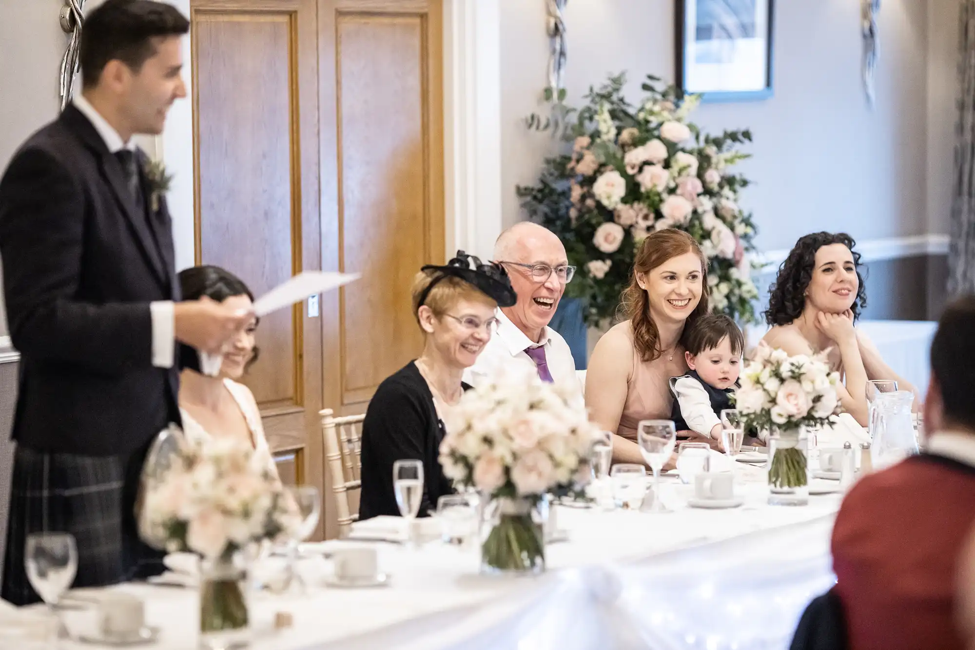 Guests seated at a wedding reception table, smiling and listening to a speech. Floral decorations are visible in the background.
