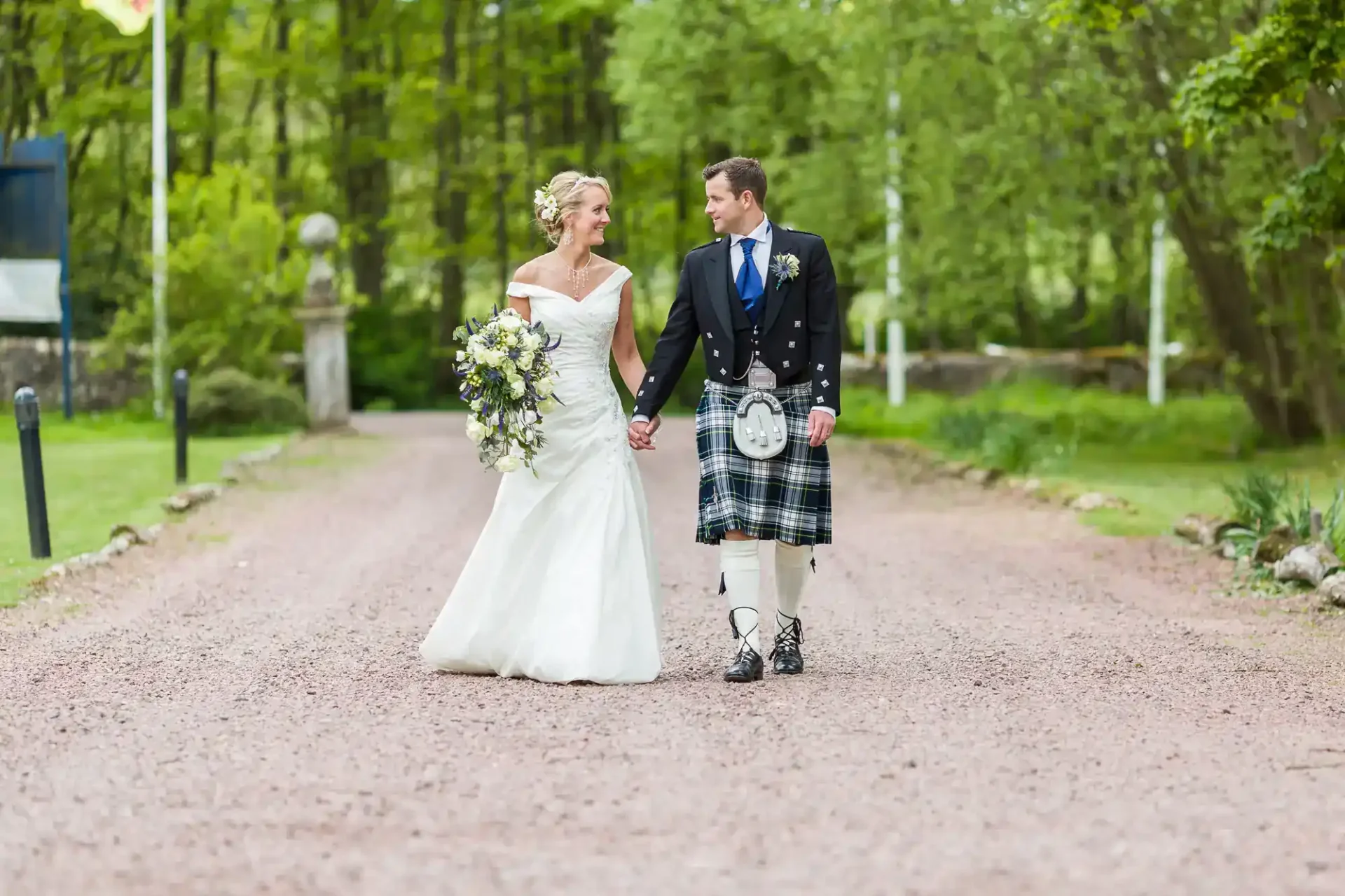 A bride in a white dress and a groom in a Scottish kilt walk hand in hand down a gravel path surrounded by greenery.