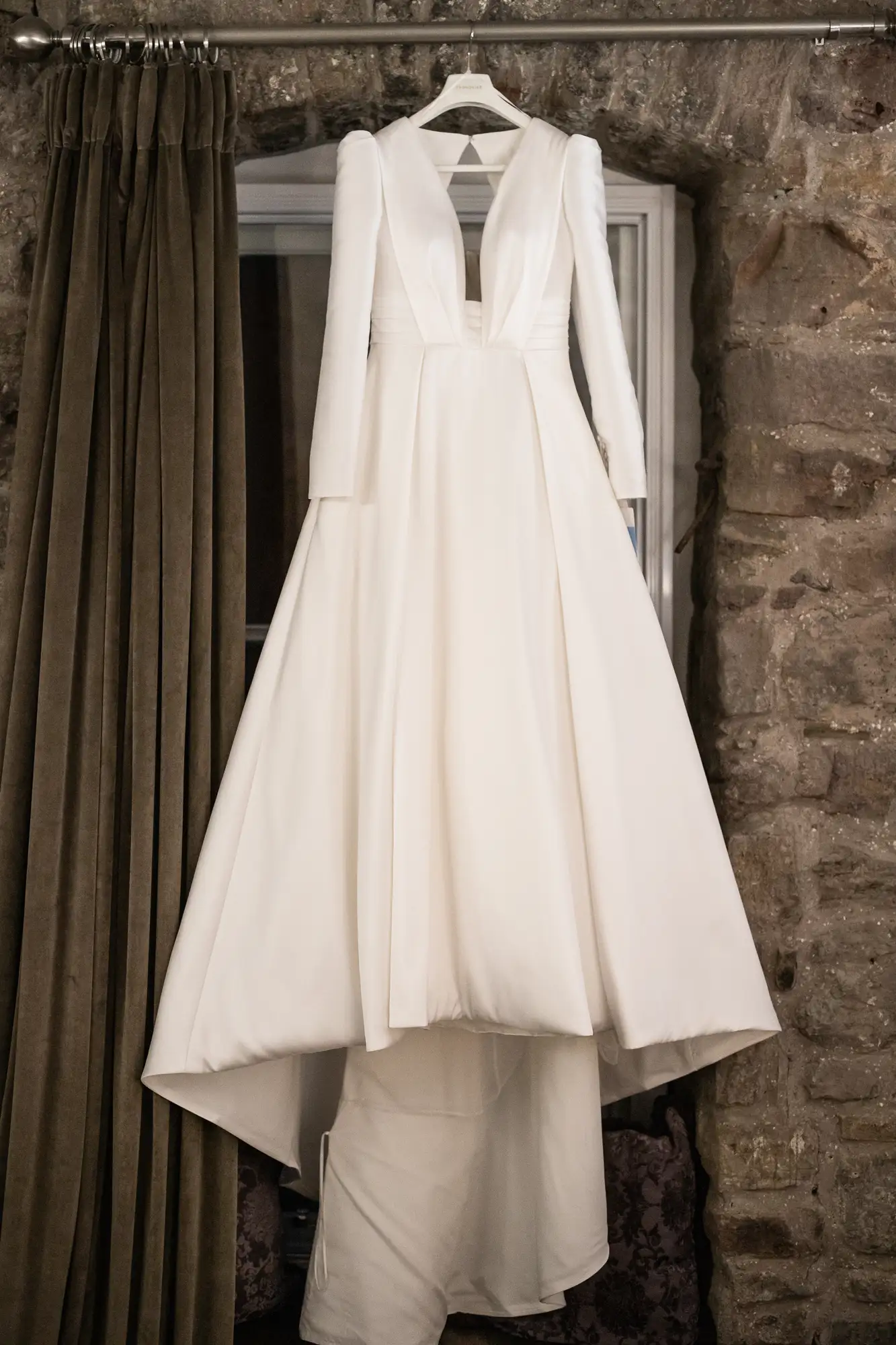 An elegant white wedding dress with long sleeves and a deep v-neckline, hanging against a rustic stone wall.