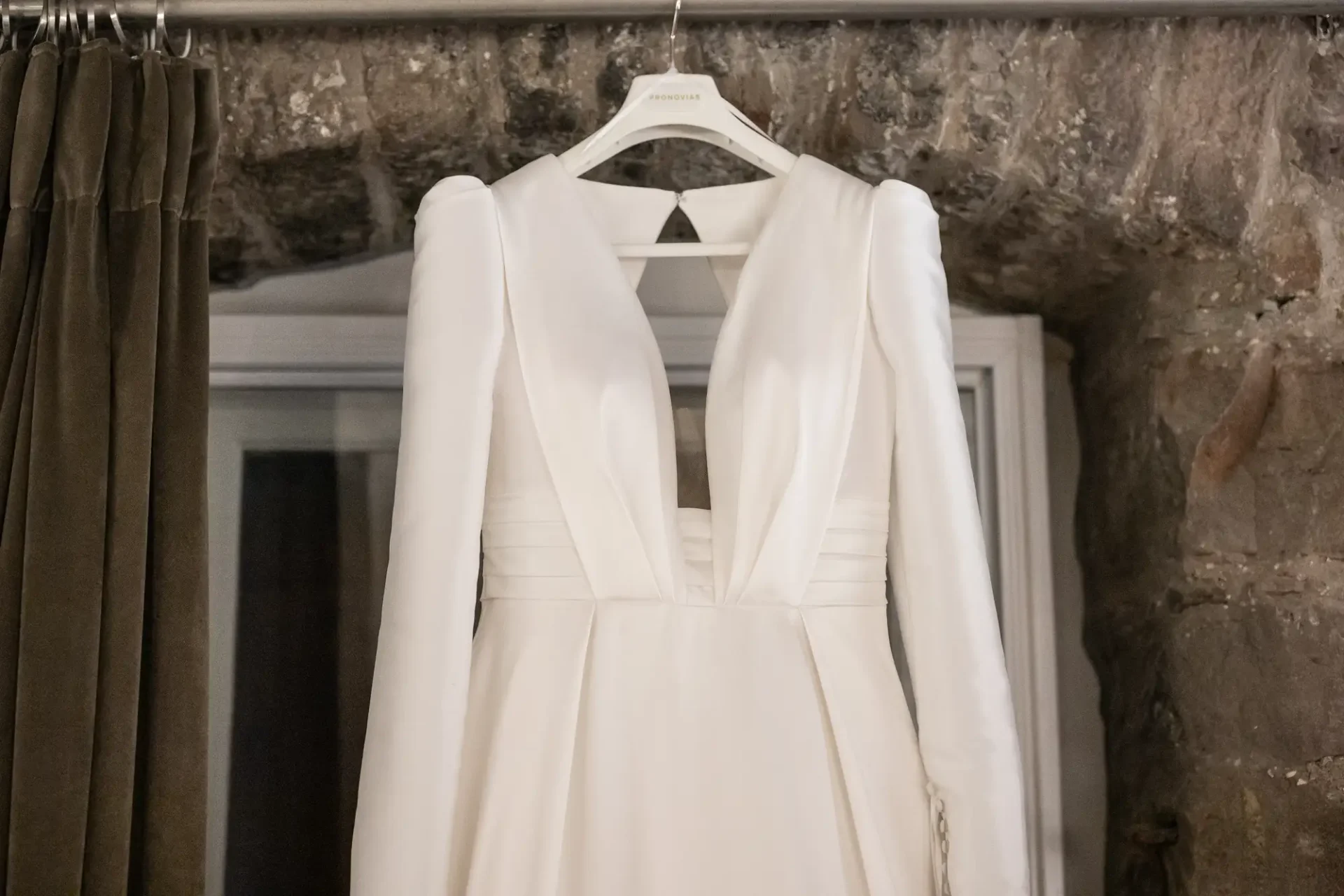 An elegant white wedding dress with long sleeves and a v-neckline, hanging on a hanger against a rustic stone wall backdrop.