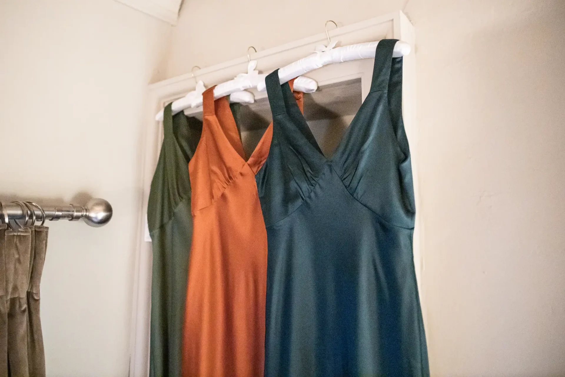 Three dresses in orange, teal, and dark green hanging on white hangers against a plain, beige wall.