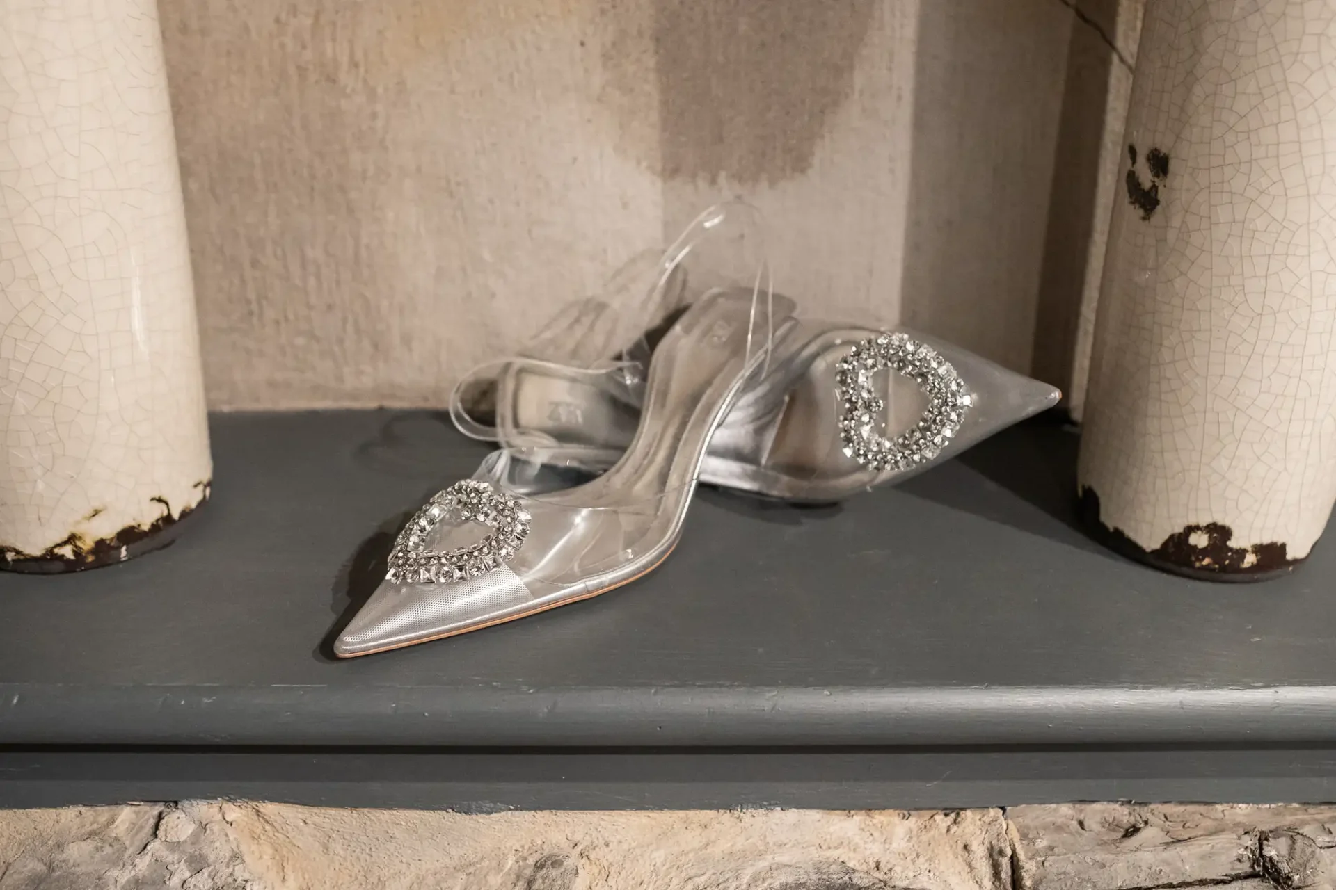 A pair of transparent high-heeled shoes with decorative crystals on the toes, displayed on a gray shelf against an aged, peeling column background.