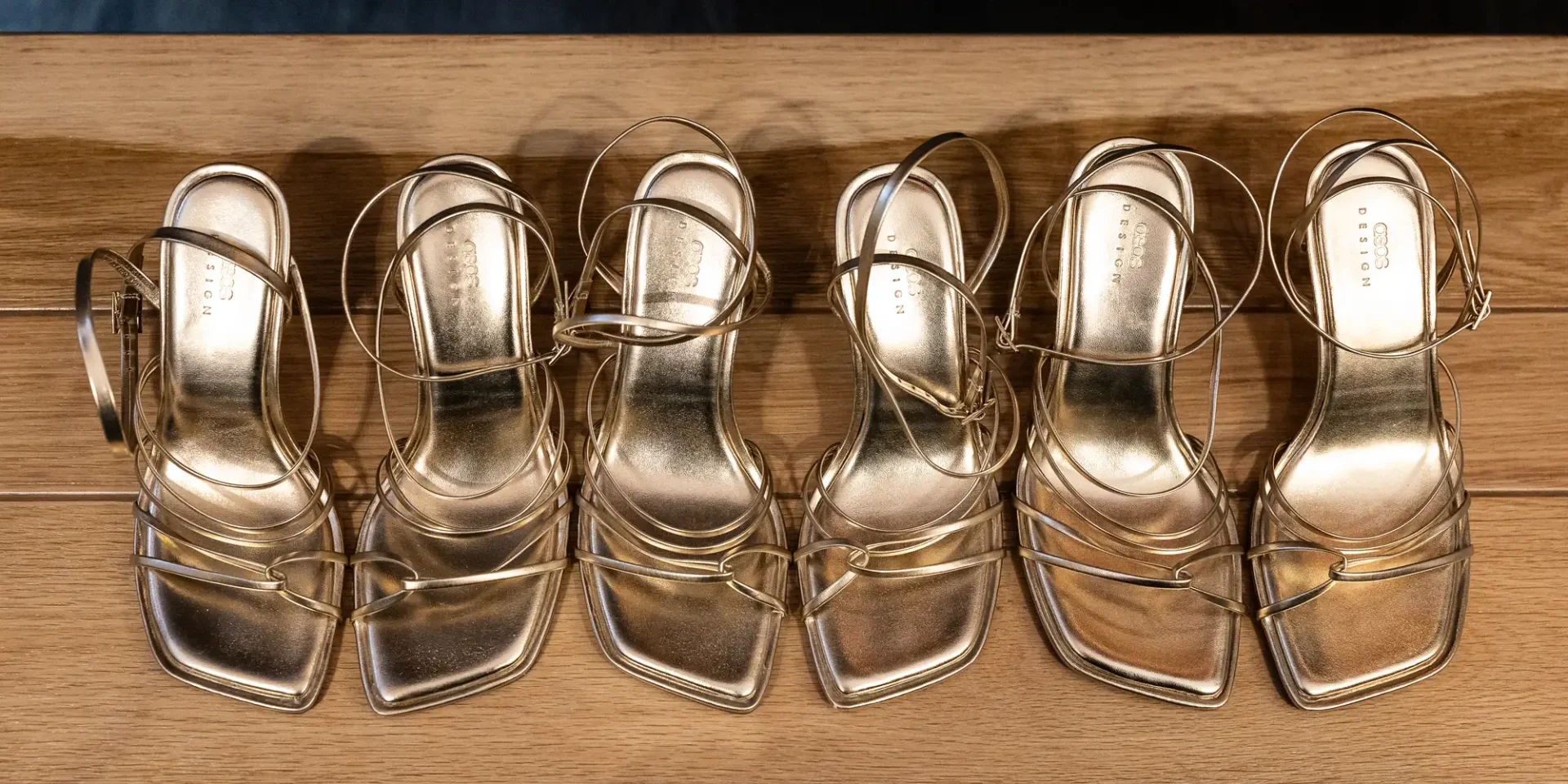 Three pairs of metallic gold high-heeled sandals with straps, displayed in a row on a wooden shelf.