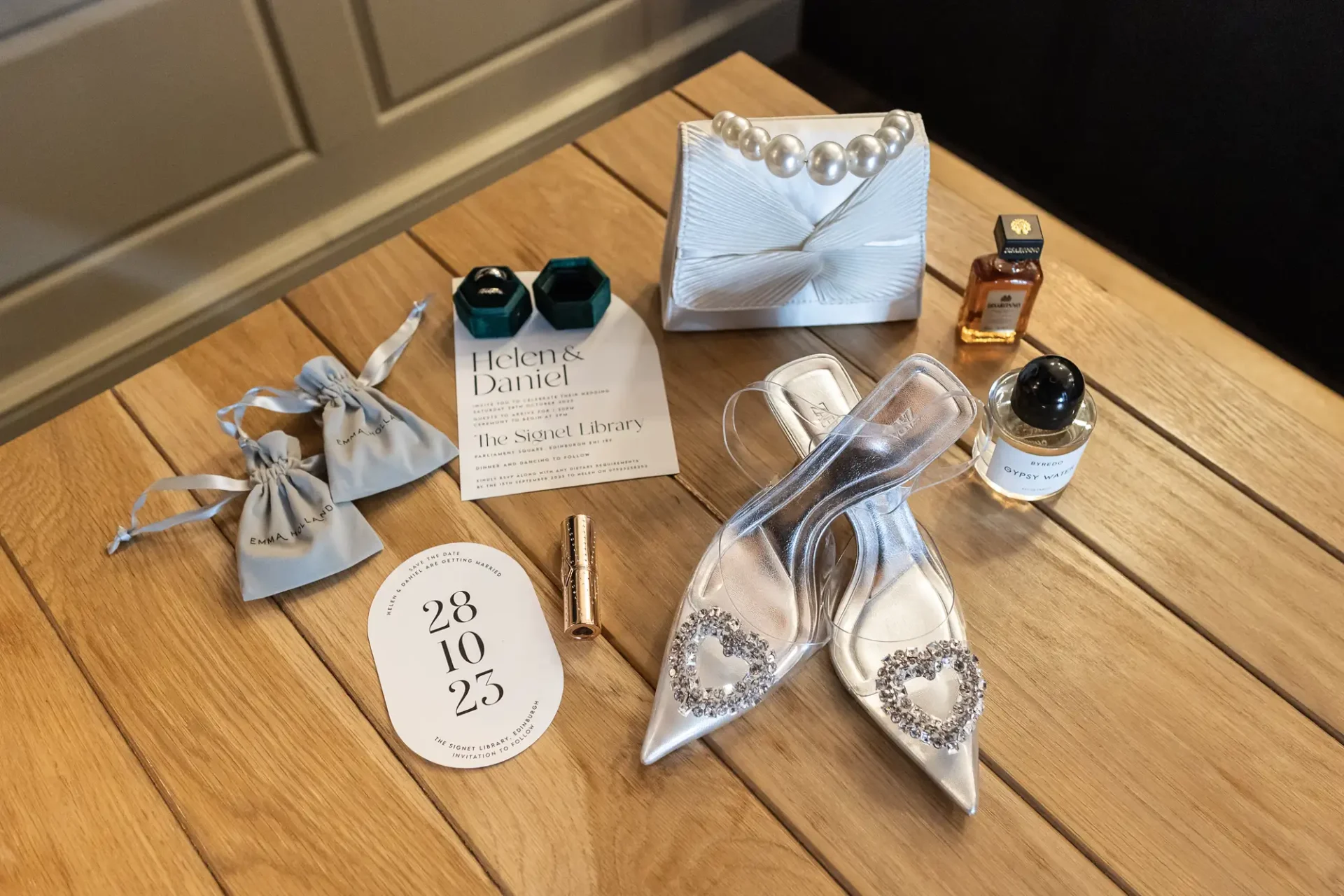 Wedding accessories arranged on a wooden floor, including bridal shoes, perfume bottles, invitation, and pearl clutch.