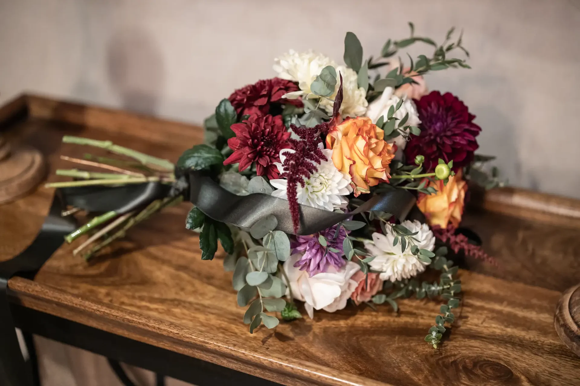 A mixed floral bouquet with dark red, orange, white, and pink flowers, accented with green foliage and a black ribbon, resting on a wooden surface.