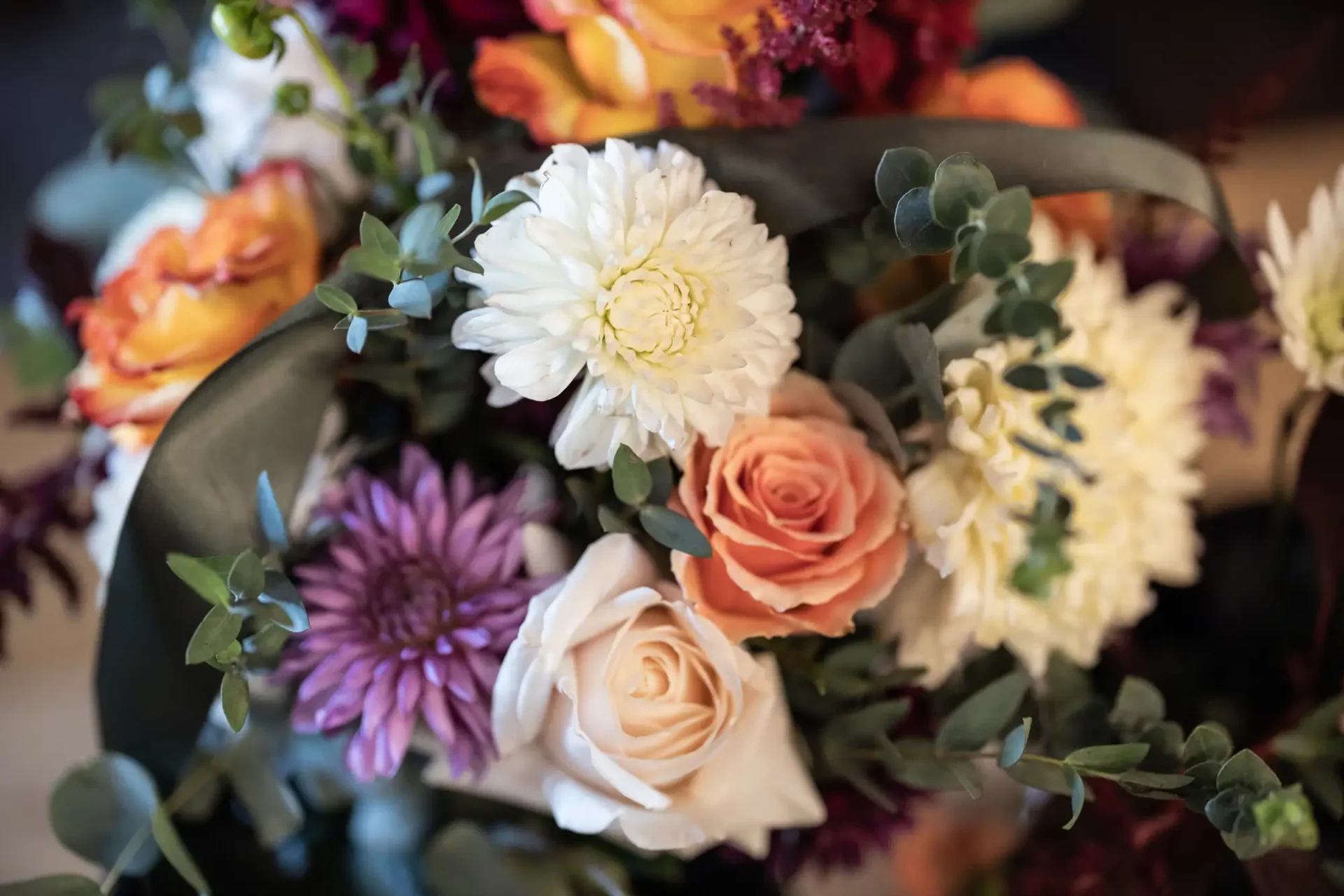 A close-up of a vibrant floral arrangement featuring white, peach, and purple flowers with greenery and ribbon accents.