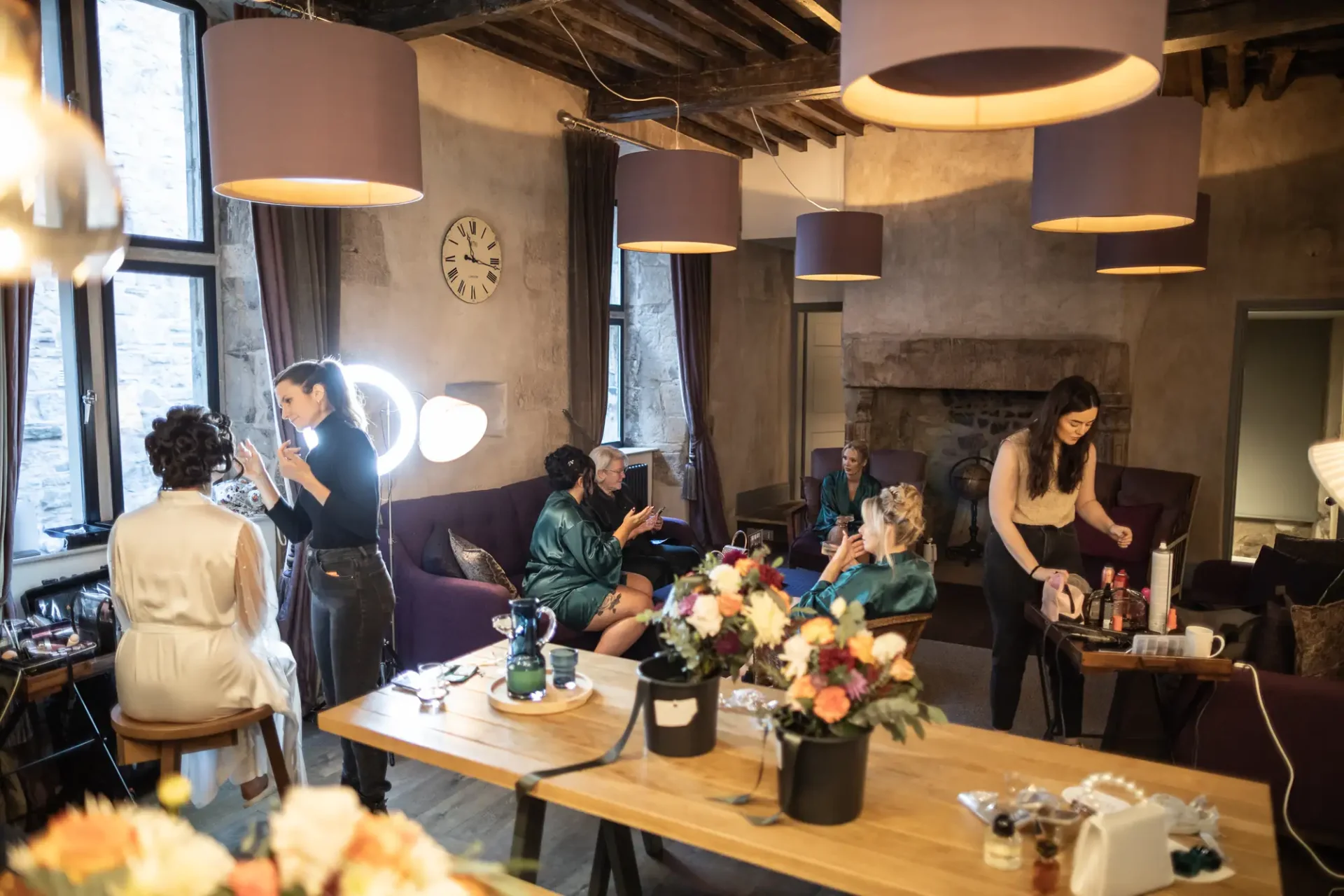 People preparing for an event in a cozy room with stone walls and modern lighting. some sit in robes getting makeup done while others engage in conversation.