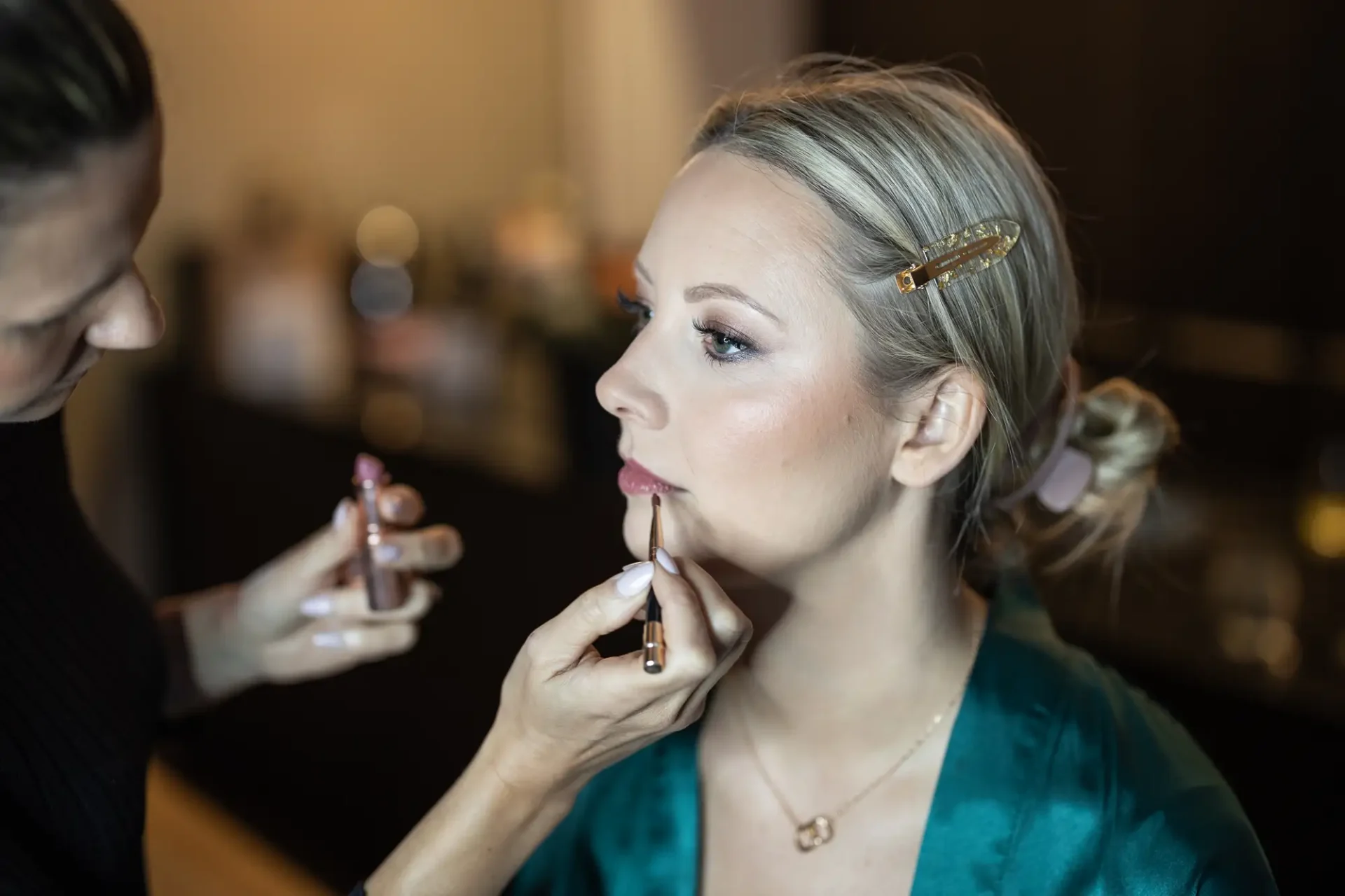 A makeup artist applies lipstick to a blonde woman in a green robe, focusing intently on perfecting her makeup.