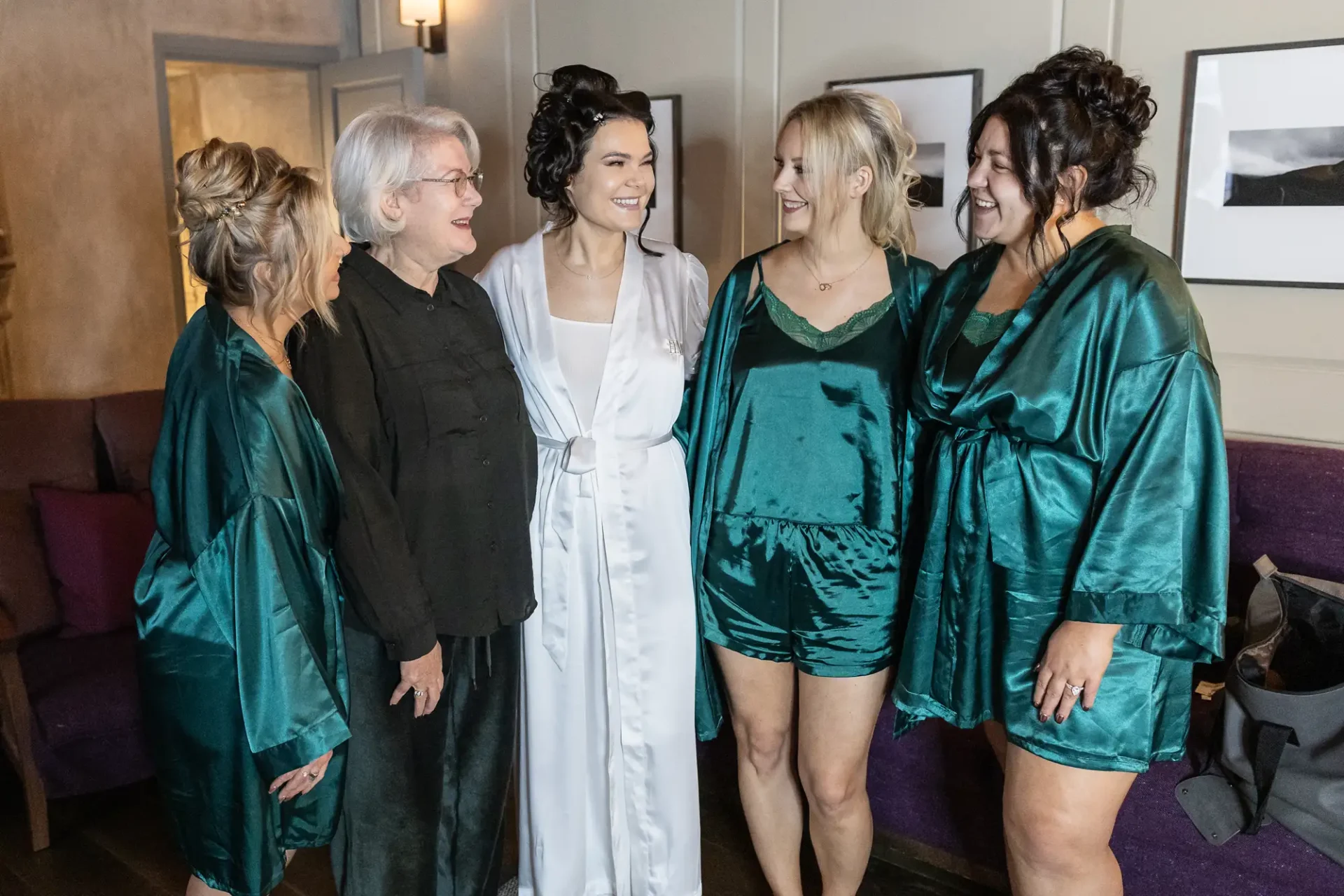Five women in robes share a joyful moment; the central woman in a white robe, presumably a bride, surrounded by others in green robes, smiling and conversing in an indoor setting.