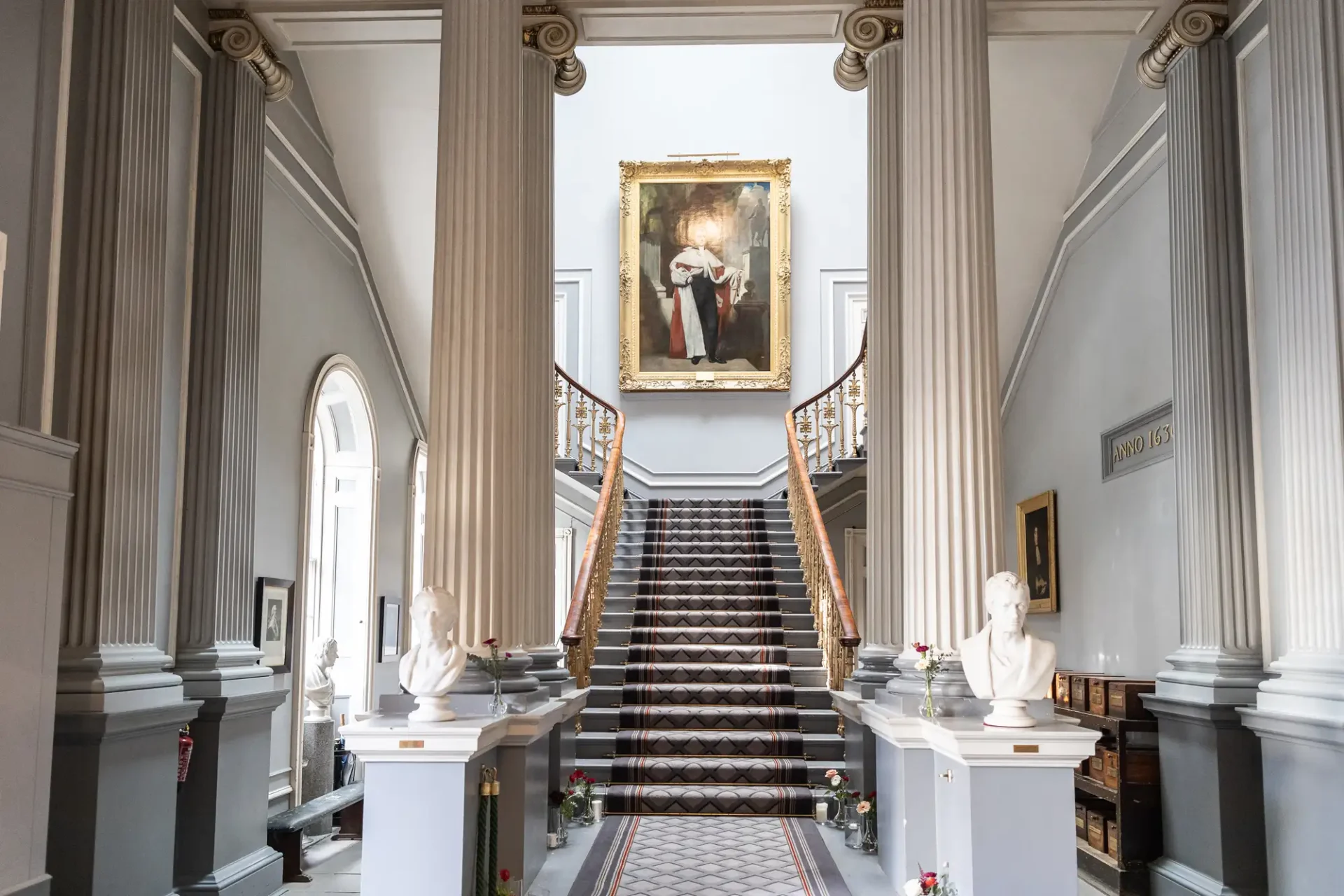 Elegant staircase in a grand hall flanked by classical columns and statues, with a large portrait hanging above.